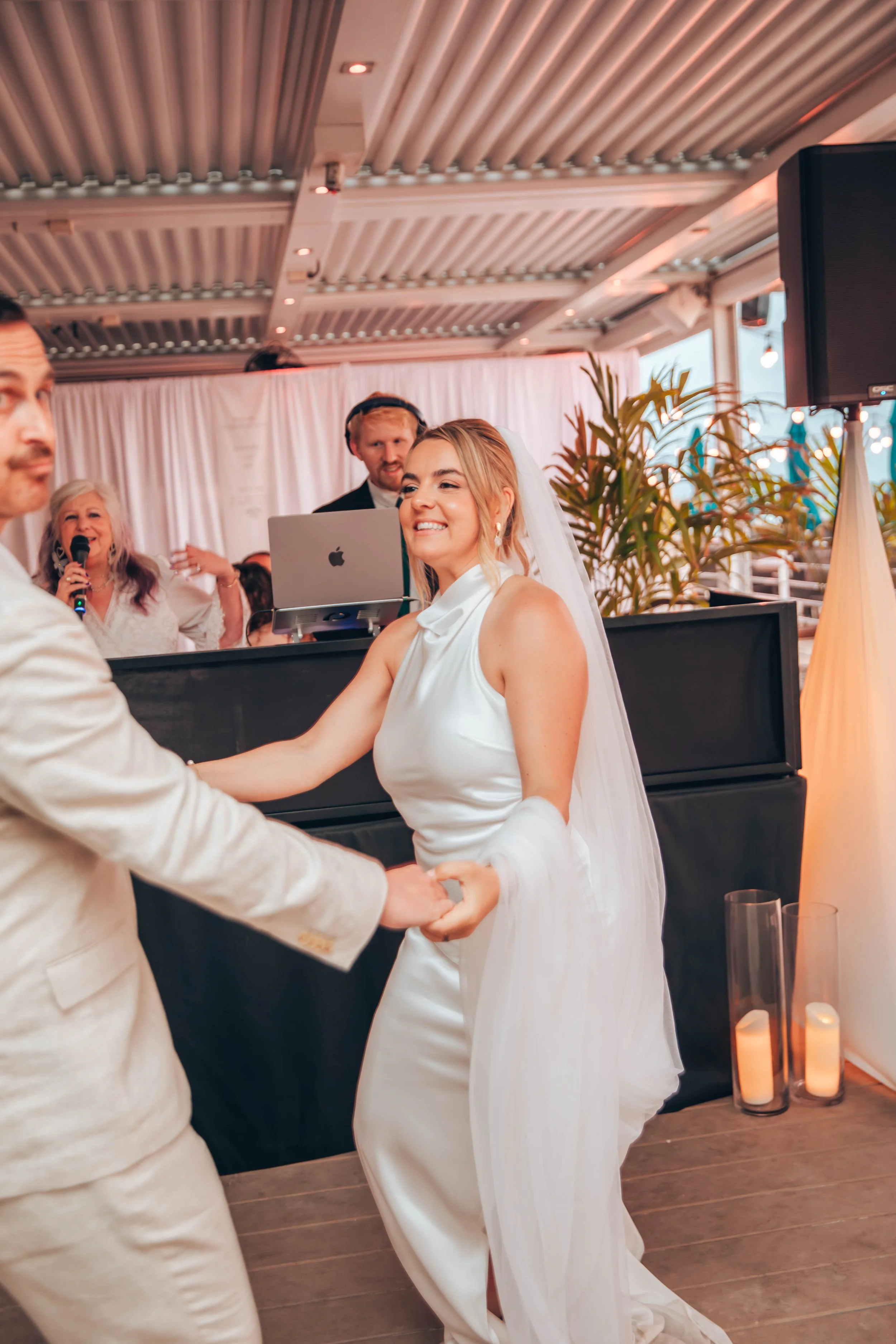 A bride and groom holding hands and smiling during their wedding ceremony, with a female officiant holding a microphone behind them and a DJ in the background, at an indoor venue decorated with candles and candles.