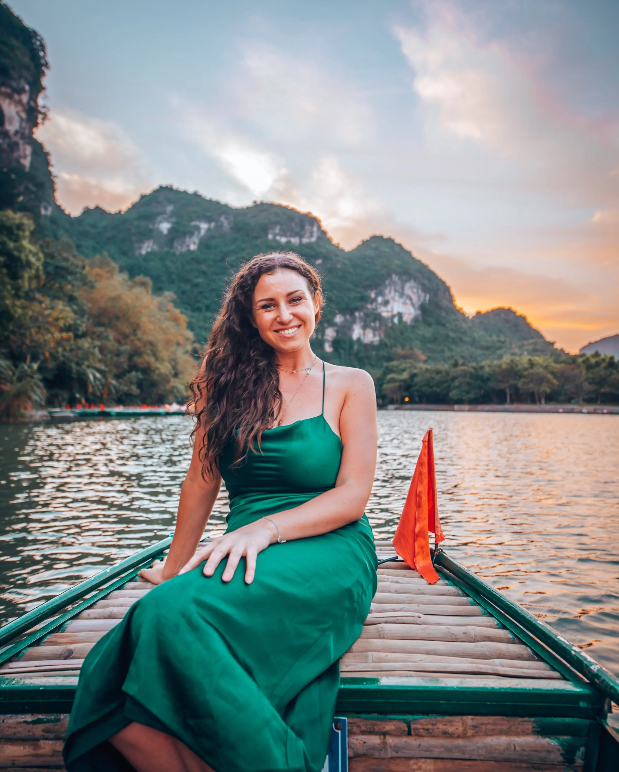 A smiling woman with long, curly dark hair sitting on a bamboo raft on a river at sunset, with green mountains in the background.