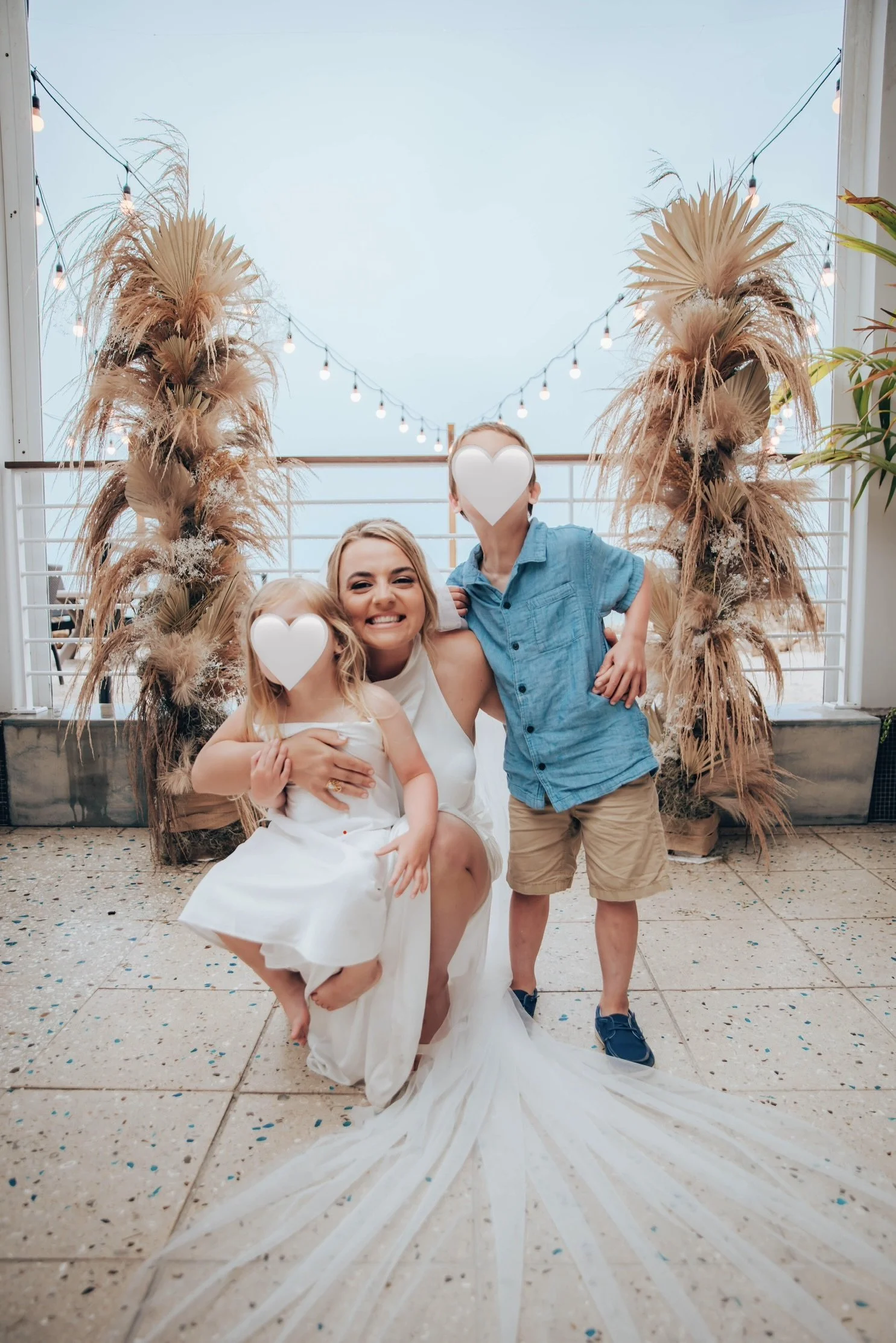 A woman in a white dress, possibly a wedding dress, is kneeling on the ground smiling. She is holding two children, one on each side. One child has heart-shaped face cover, and the other is a boy wearing a blue shirt and tan shorts. They are under an
