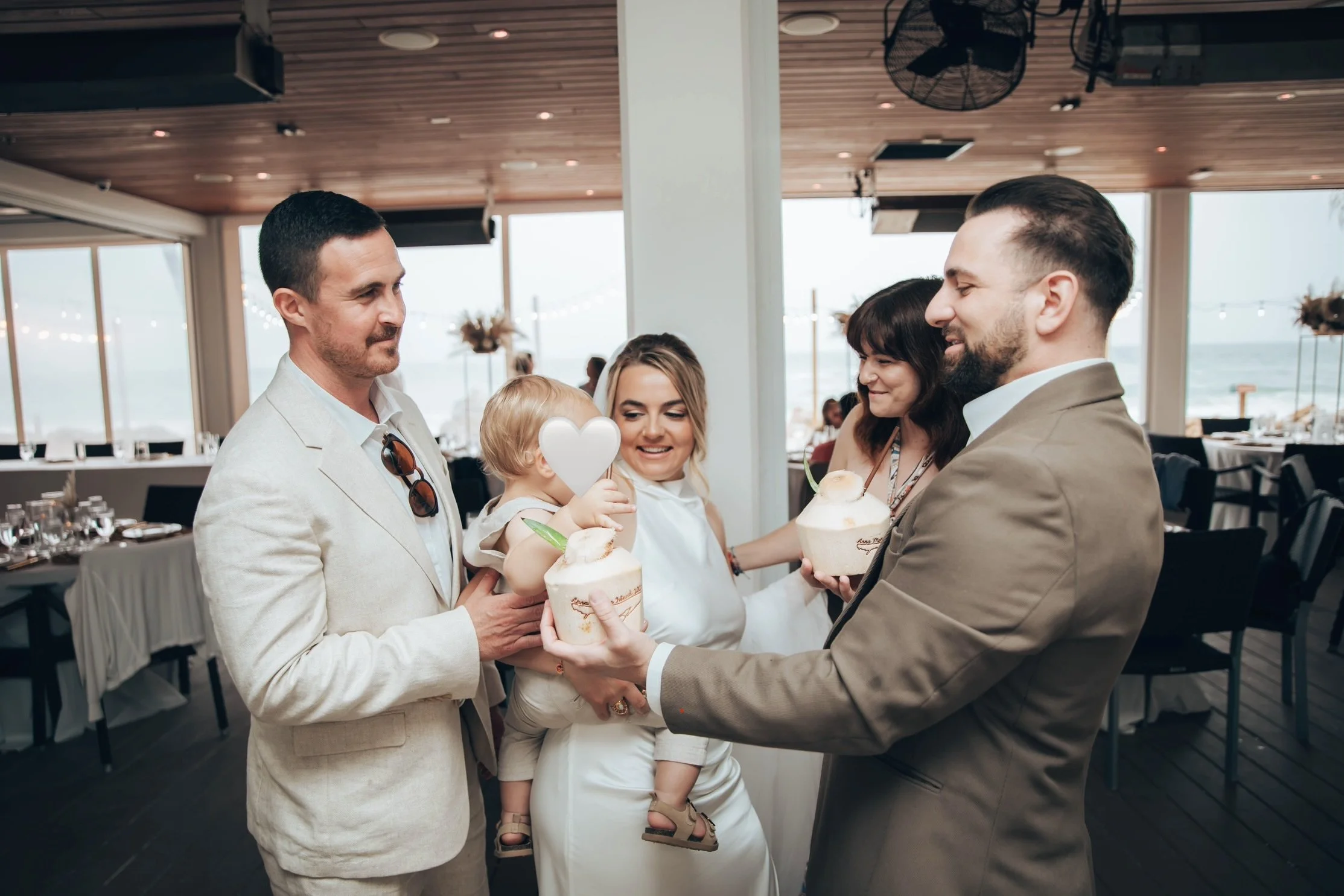 A group of five people, three adults and two children, standing inside a restaurant near a window overlooking a beach, holding coconuts with straws, celebrating at a social gathering.