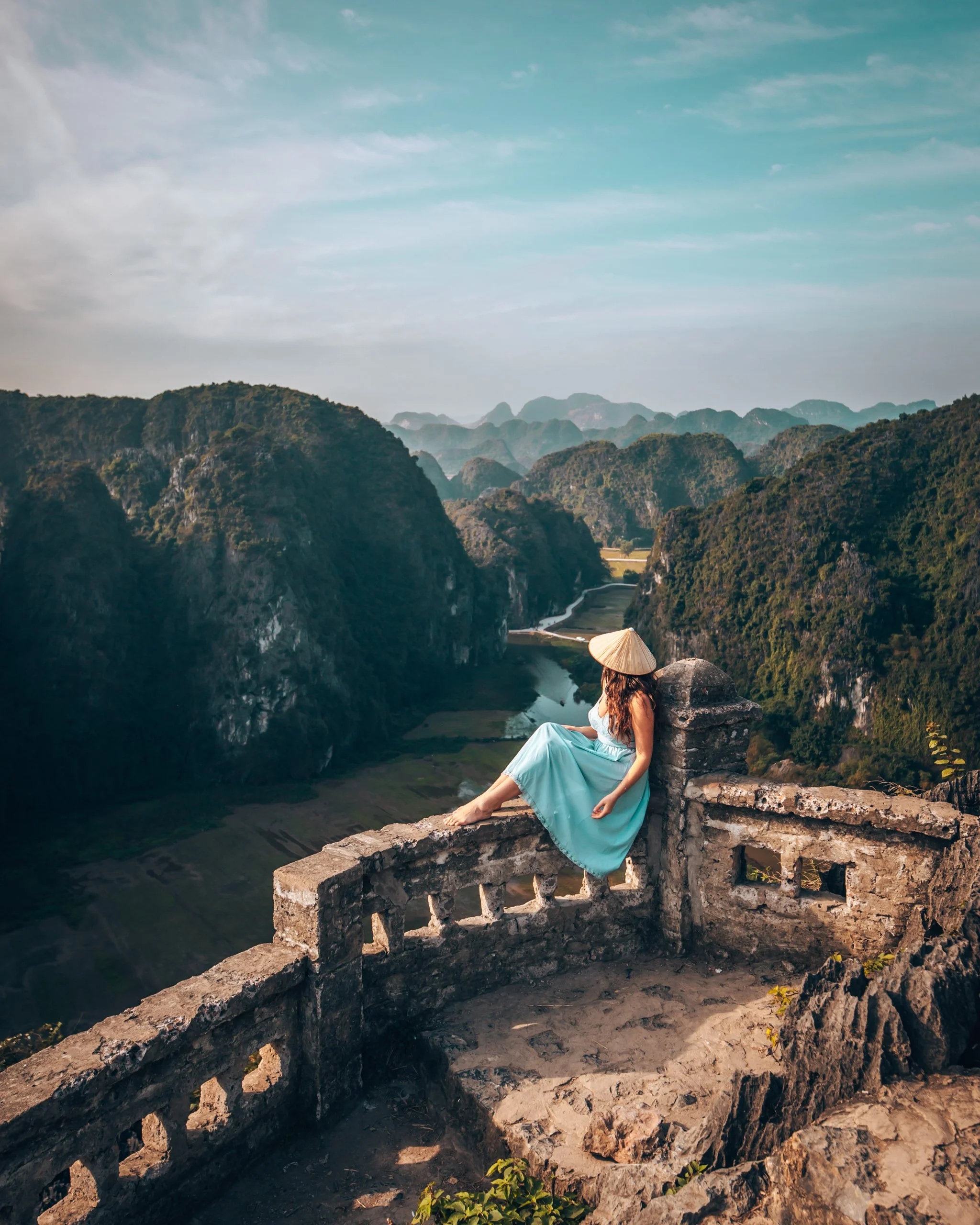 A woman sitting on a stone railing overlooking lush green mountains and a river beneath a partly cloudy sky. She is wearing a light blue dress and a traditional Asian conical hat.
