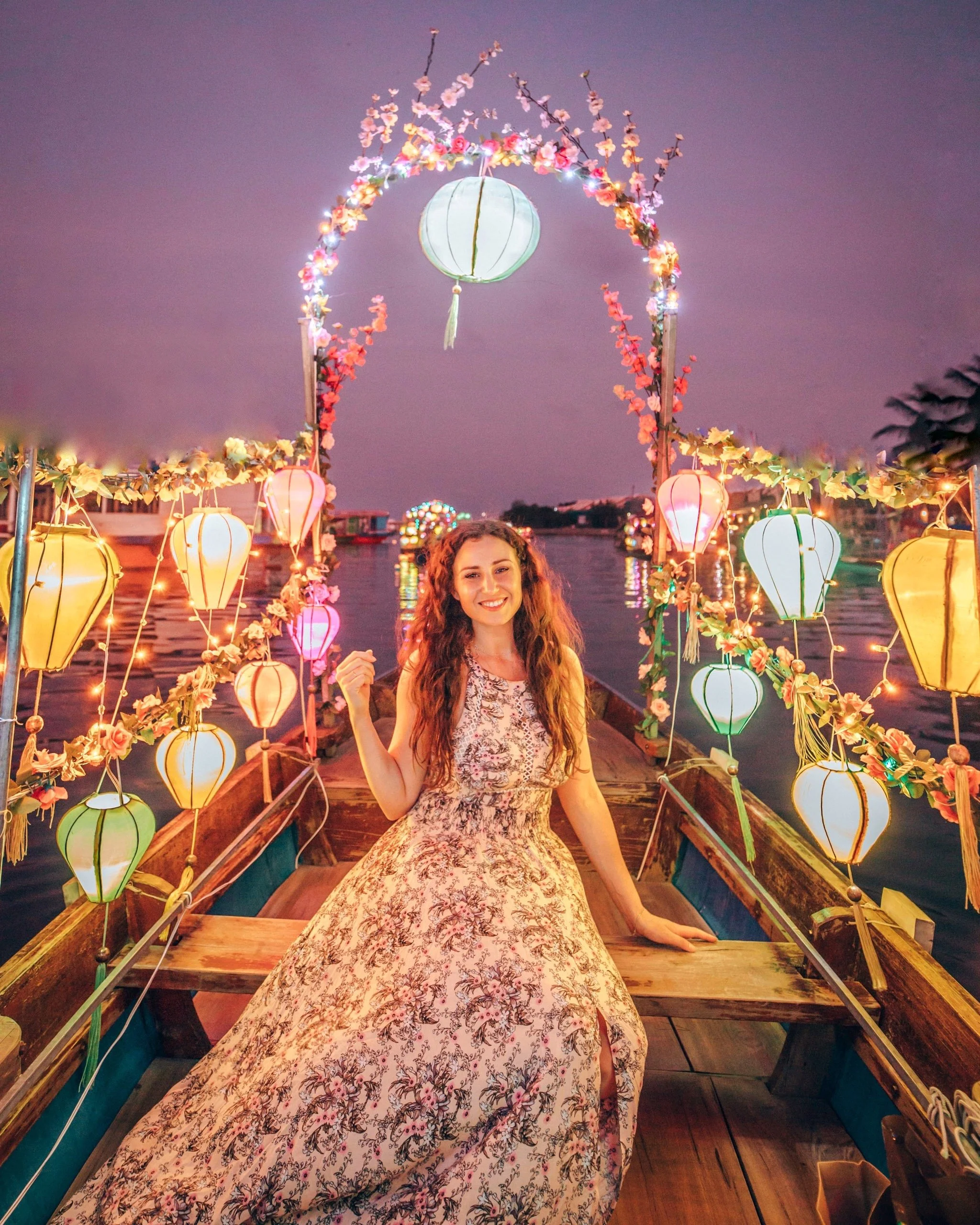 A young woman in a floral dress is sitting in a decorated boat at dusk, surrounded by colorful lanterns and string lights, with a waterway and ferris wheel in the background.