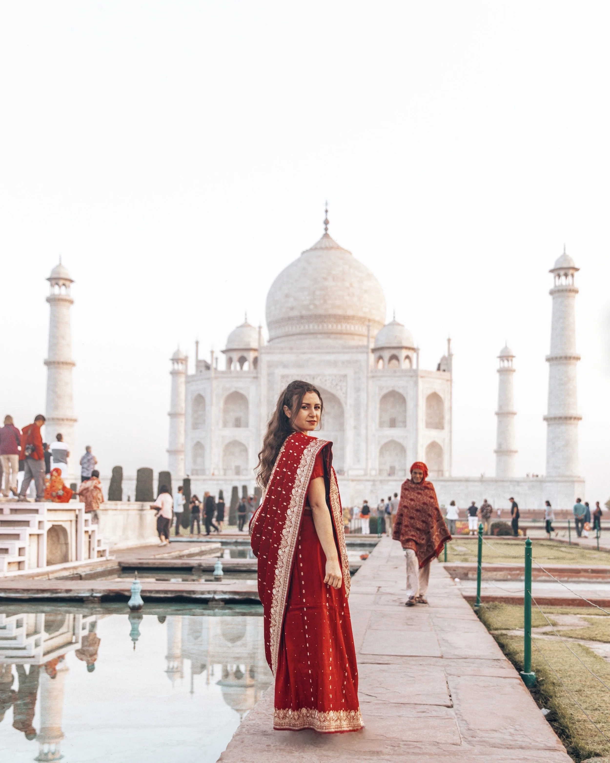 A young woman in a red traditional Indian saree stands near the Taj Mahal in India with the monument's white marble domes and minarets in the background. There are other visitors around her, and the scene is outdoors with clear sky.