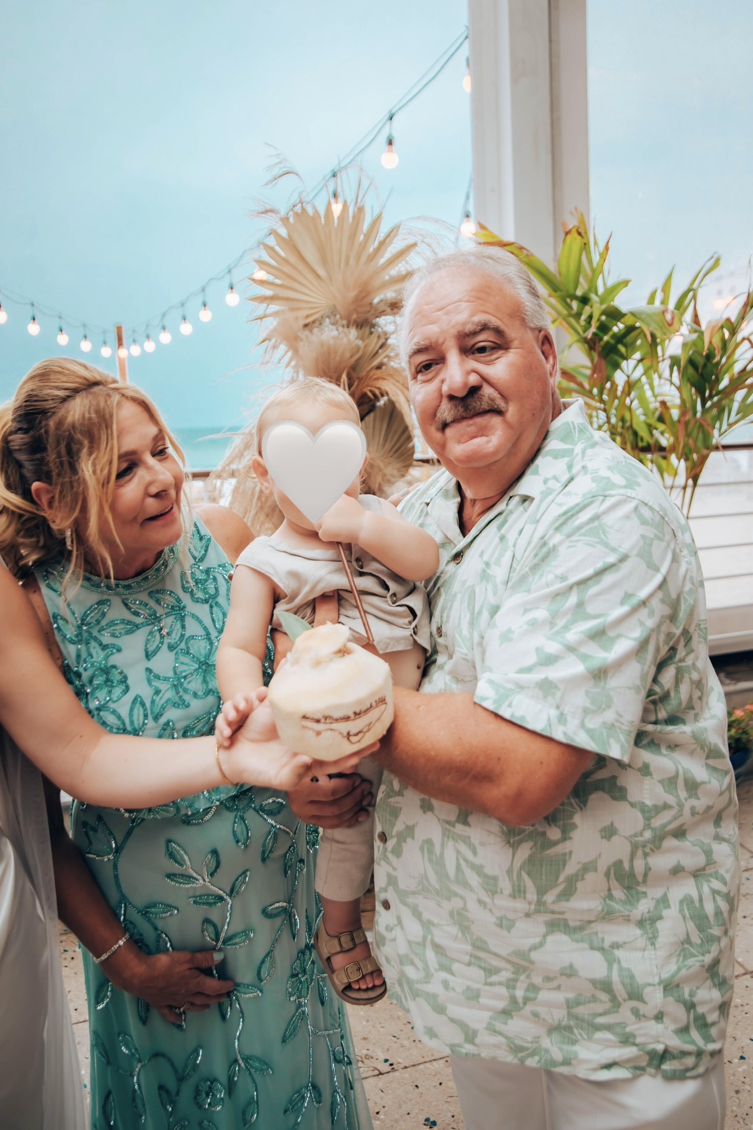 A group of people celebrating, including a woman in a turquoise dress holding a young child with a heart shape balloon covering their face, and an older man in a green patterned shirt. They are outdoors with string lights and tropical plants in the b