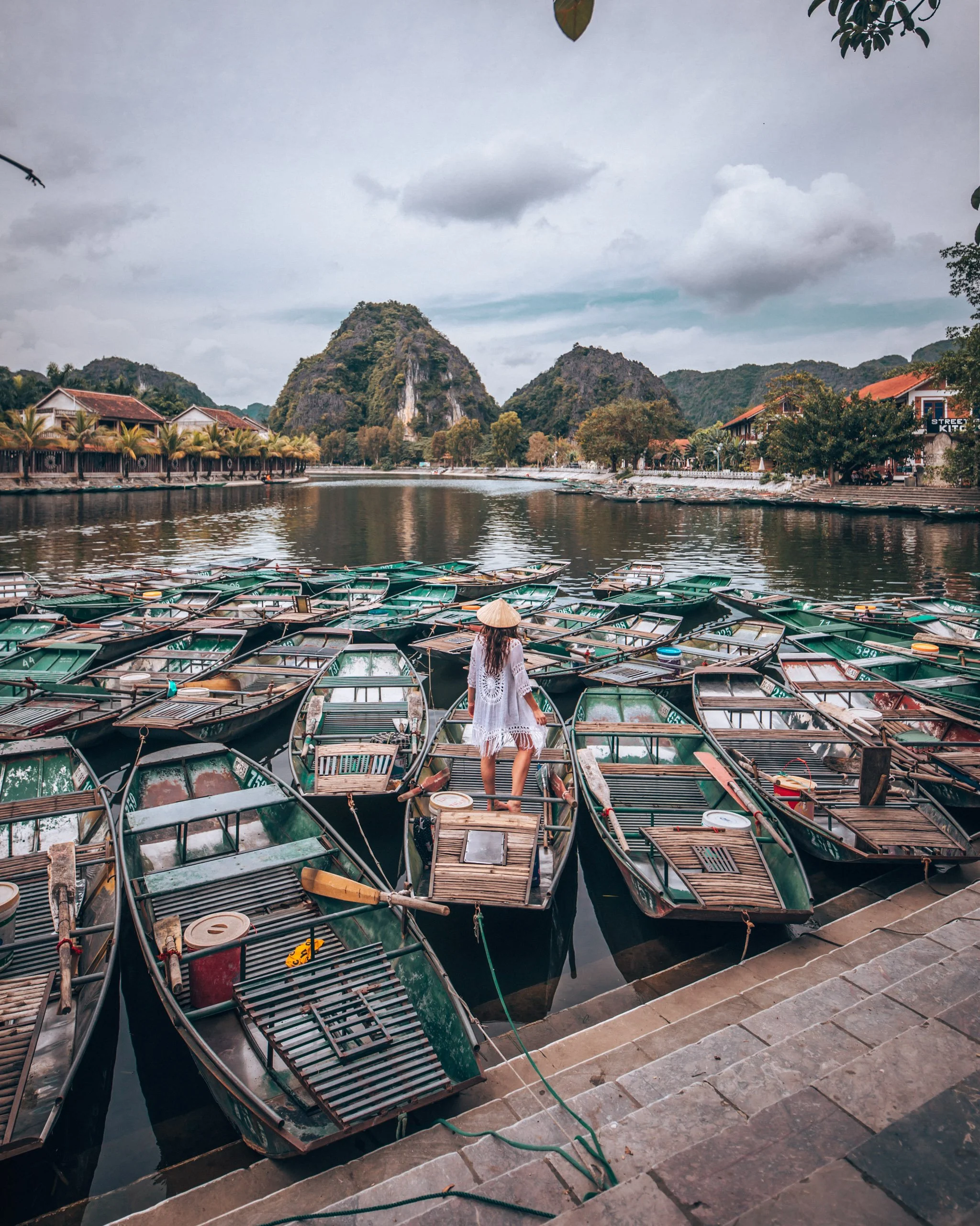 A lakeside scene with boats docked along a quay, a woman in a white dress on the dock, buildings with red roofs, and lush green mountains in the background under cloudy skies.