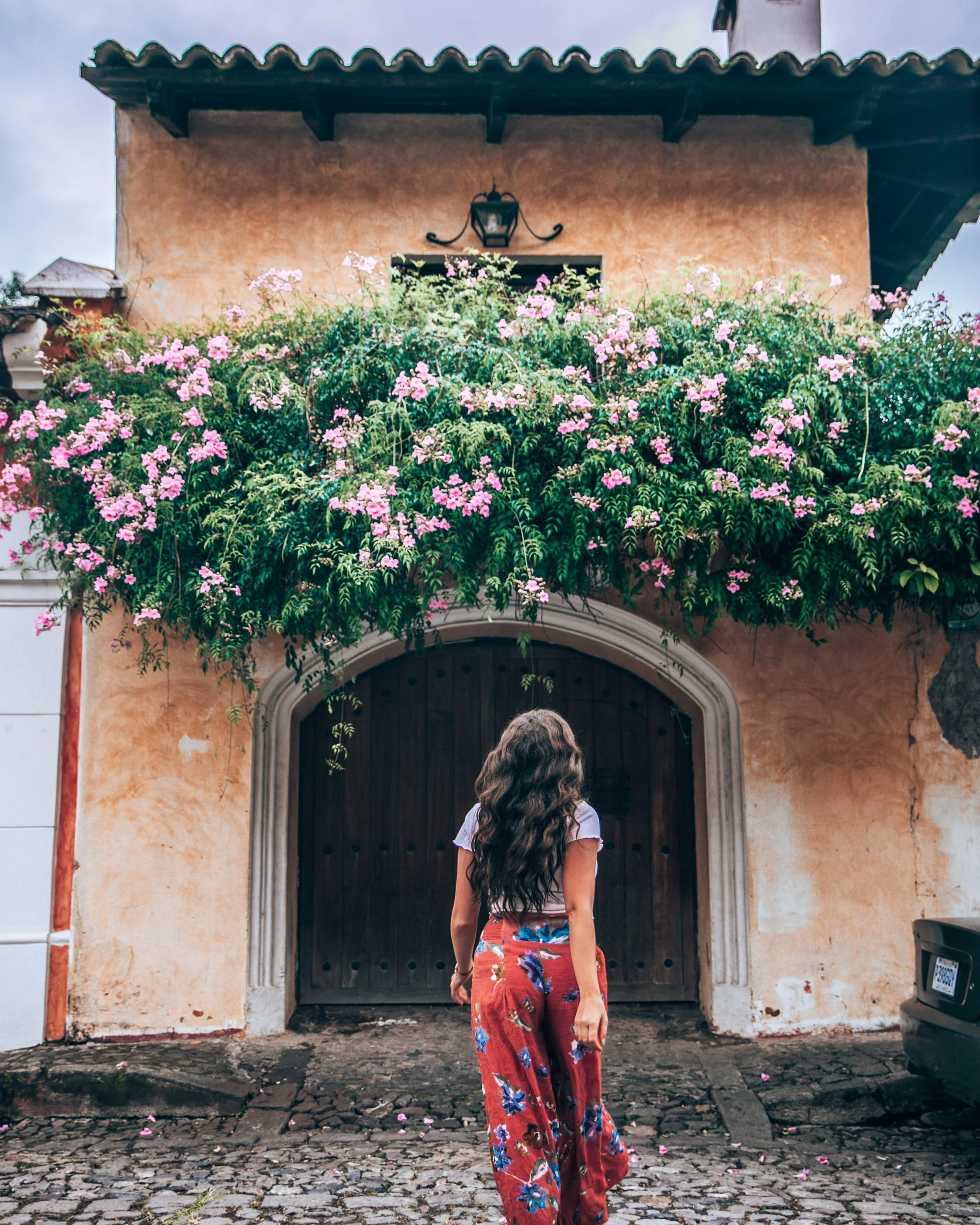A person with long, curly hair is standing in front of a large, dark wooden gate framed by an arched architecture. The individual is wearing red pants with a colorful floral pattern and a white top. Above the gate, there is a flower arrangement with 