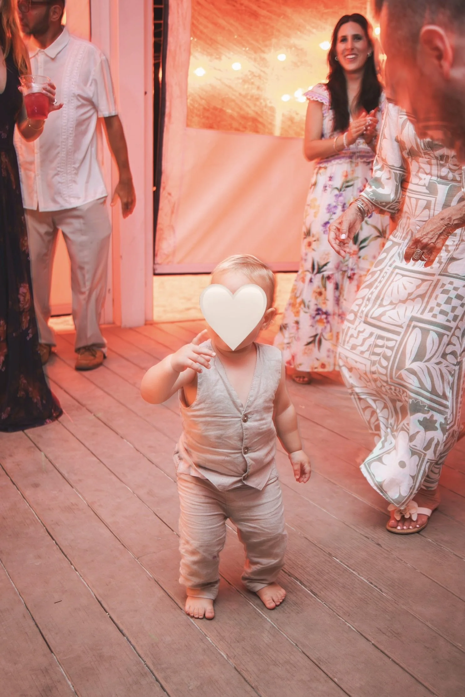 A young child with a heart emoji covering their face standing on a wooden floor at a social gathering, surrounded by adults wearing colorful clothing, some holding drinks, with warm lighting and a festive atmosphere.