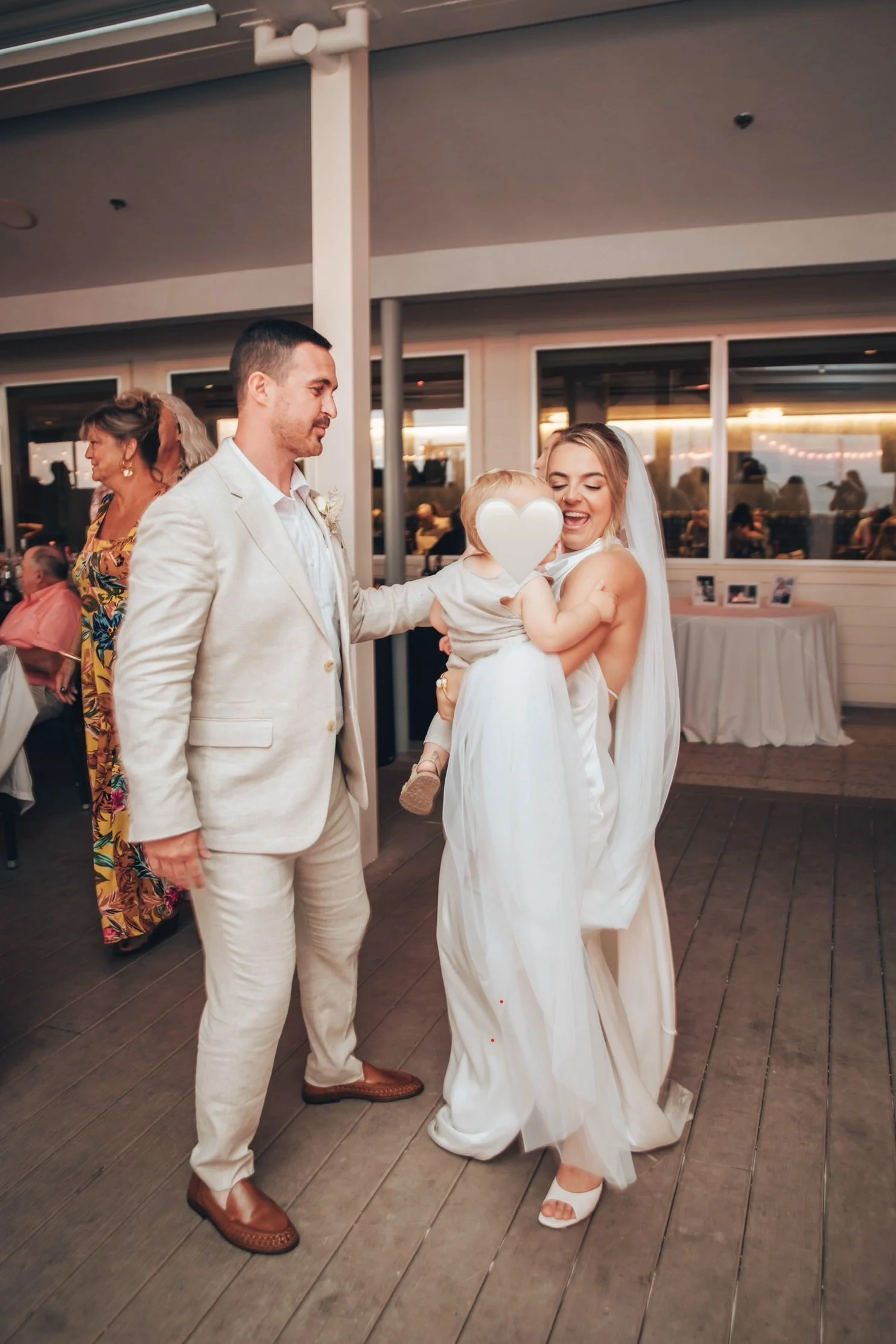 A woman in a white wedding dress is holding a young child with a heart shape superimposed over the child's face. She is smiling and looking at the child. A man in a light-colored suit is standing next to her, watching the scene. Other people are in t