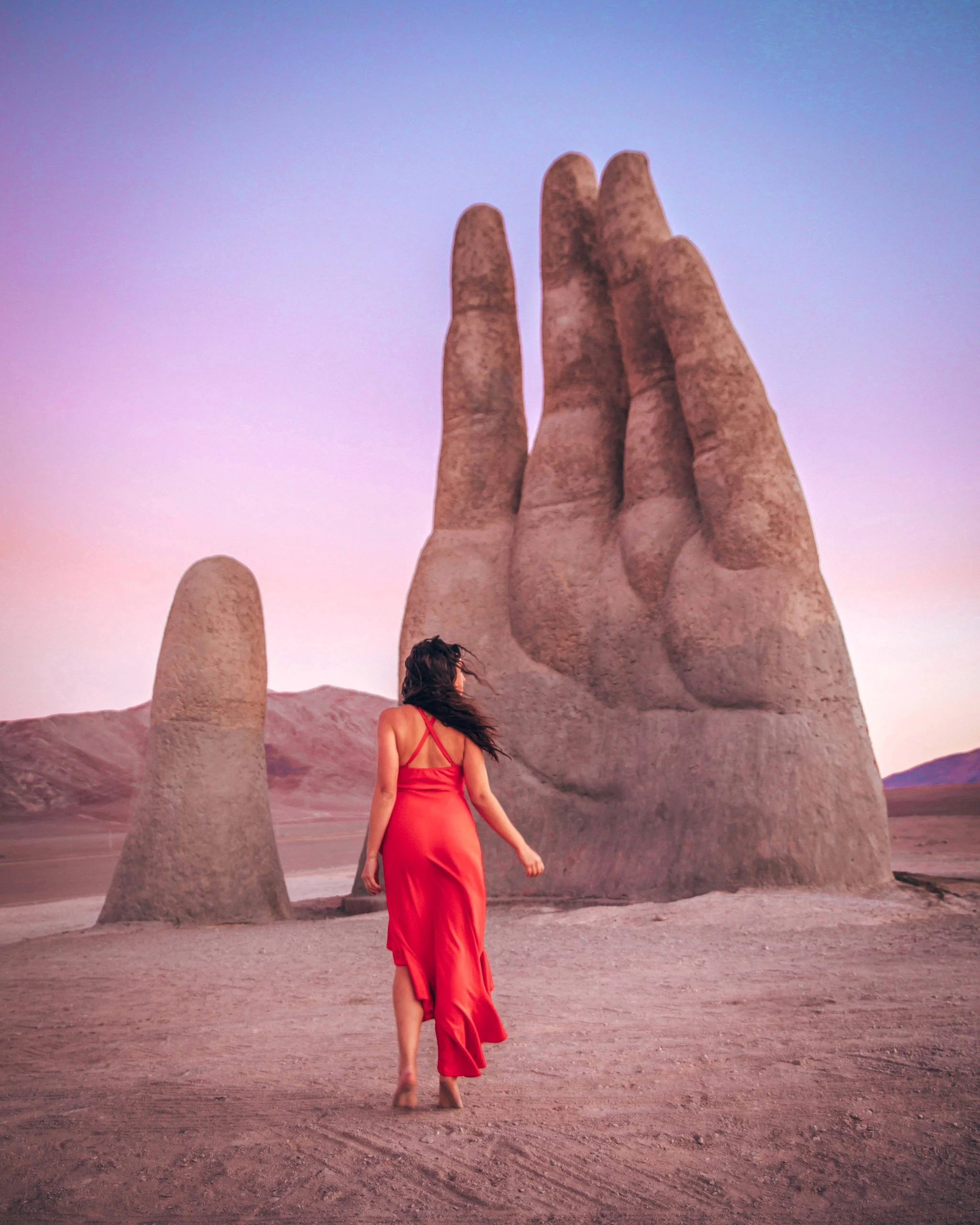 A woman in a red dress walking on a desert landscape towards a large stone sculpture of a hand with three fingers extended, set against a pastel-colored sky at sunset.