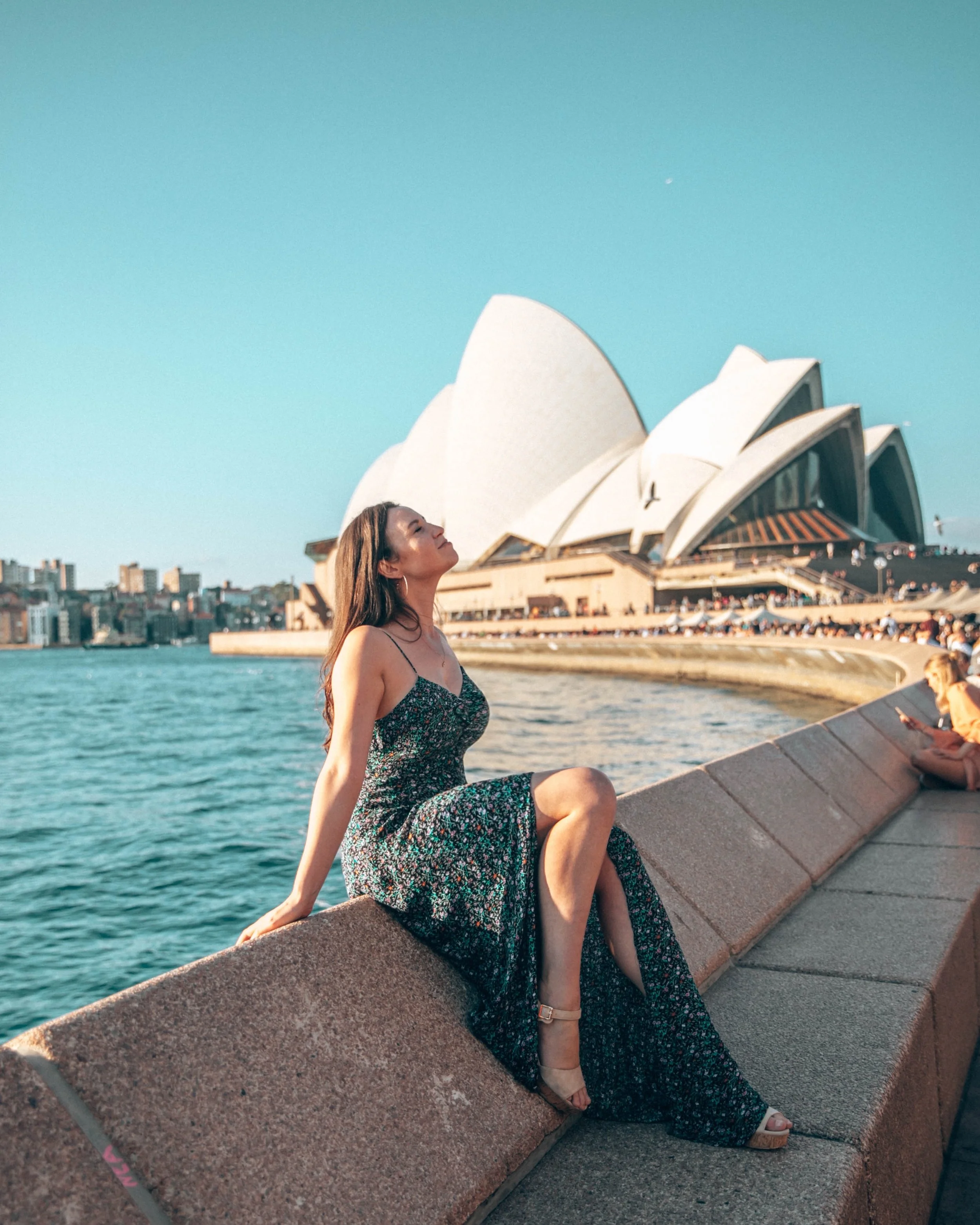 A woman wearing a floral dress sitting on a stone ledge near the water with the Sydney Opera House in the background.