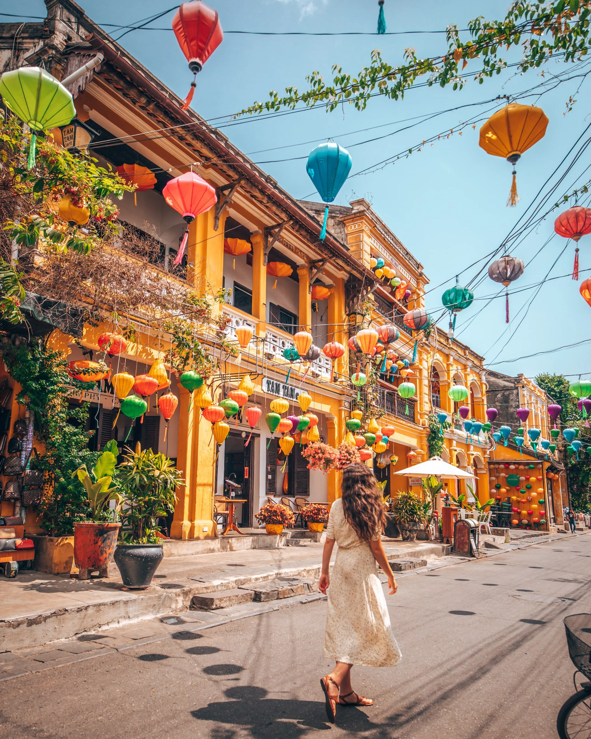 A woman in a long floral dress walking down a street decorated with colorful lanterns hanging above. The street has yellow buildings with balconies, green plants, and outdoor seating under umbrellas.