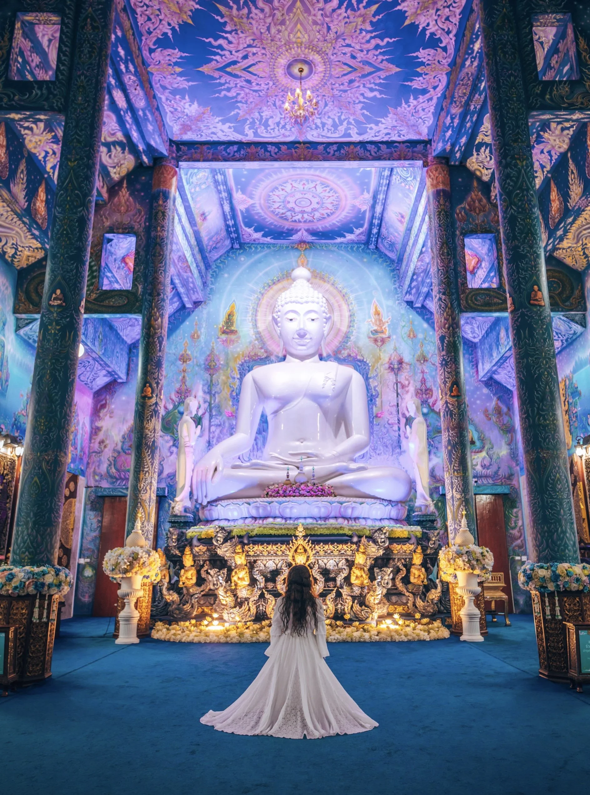 A woman in a white dress kneels in prayer before a large white Buddha statue inside a decorated temple with ornate blue and purple ceiling and pillars.