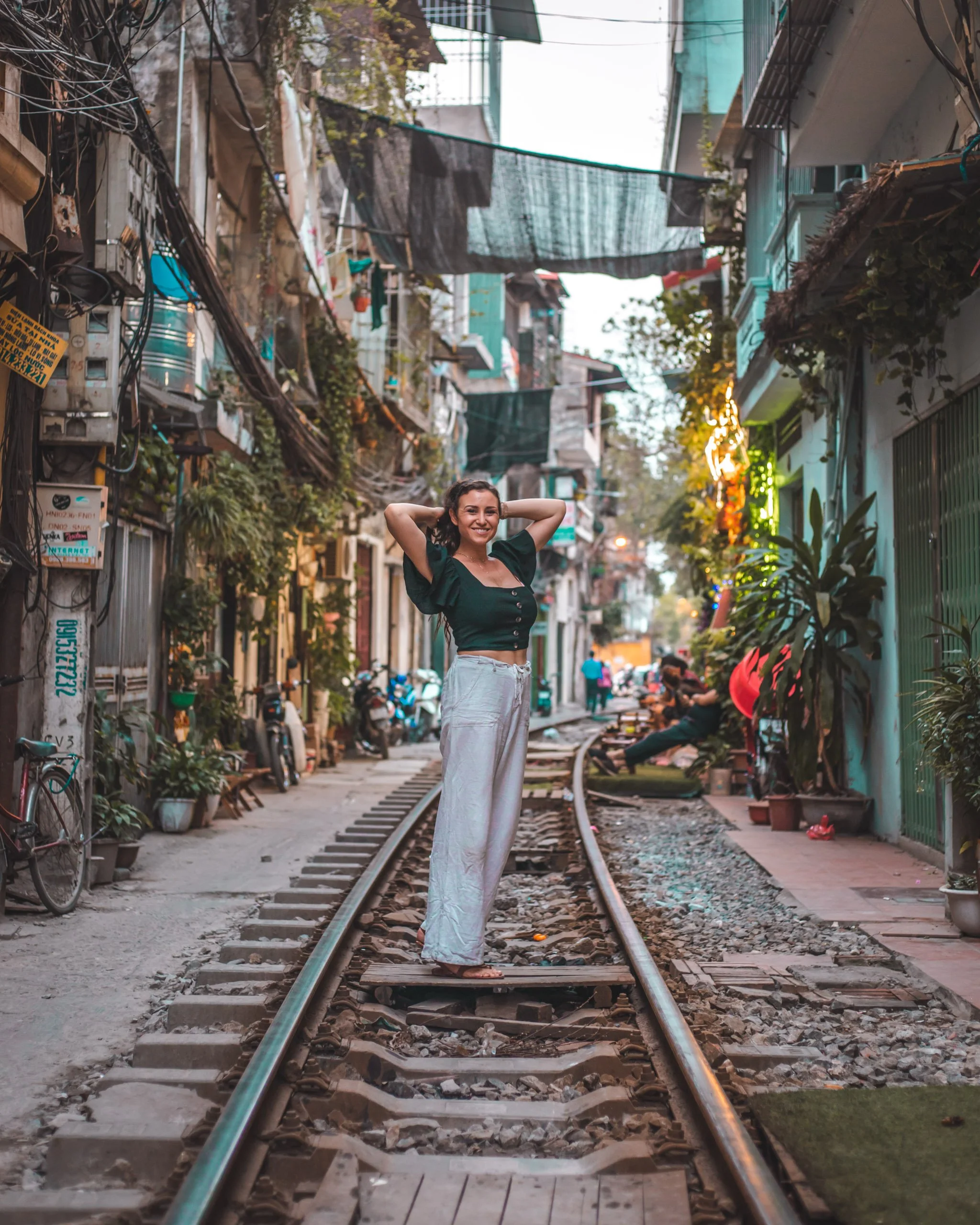 A woman standing on a railway track in an urban alleyway, smiling and posing with her hands behind her head. The alley is lined with plants, electrical wires, and buildings with balconies, and appears to be in a residential area.