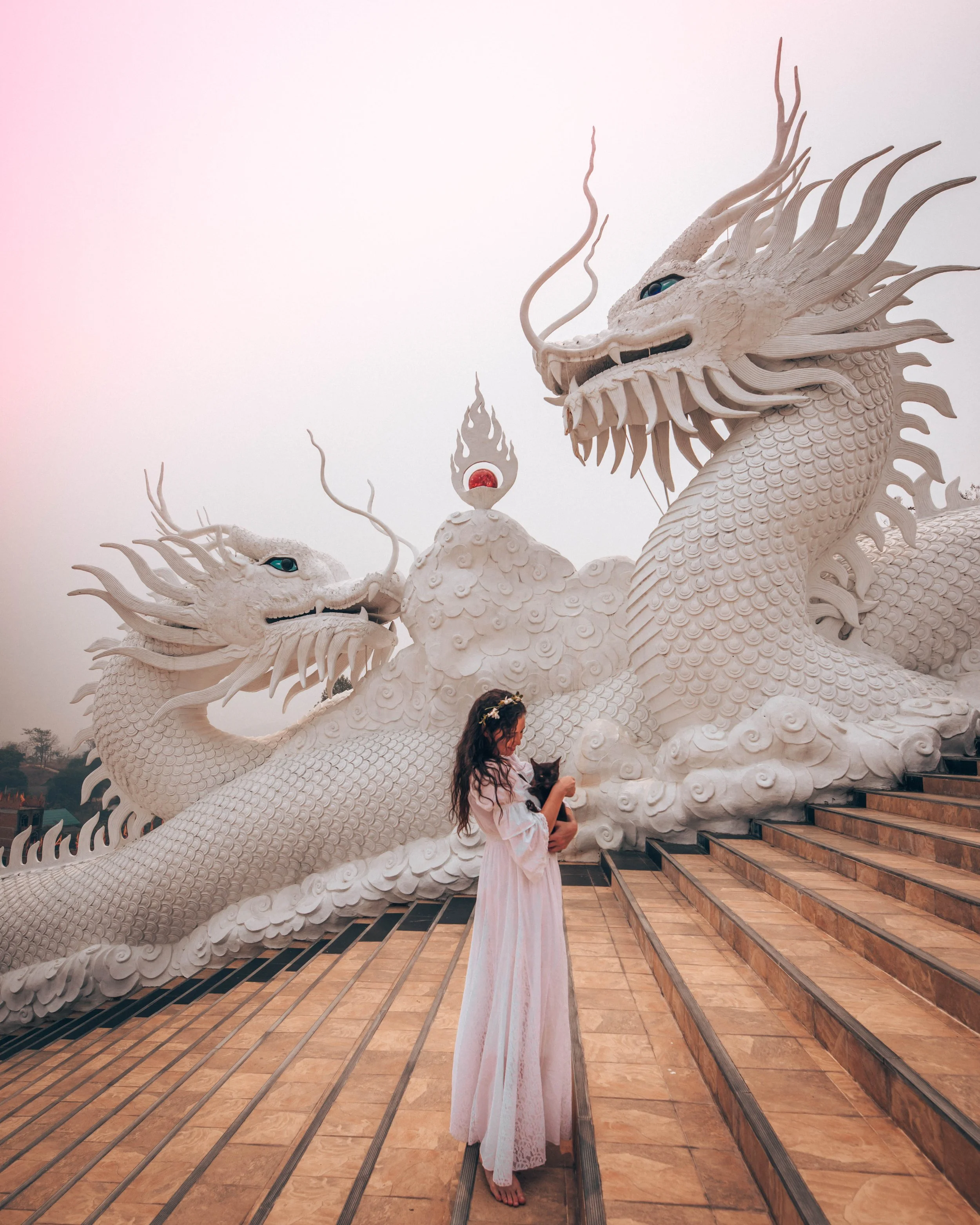 A woman in a light pink dress holding a small black dog stands at the bottom of a staircase in front of a large white dragon sculpture with intricate details and blue eyes.