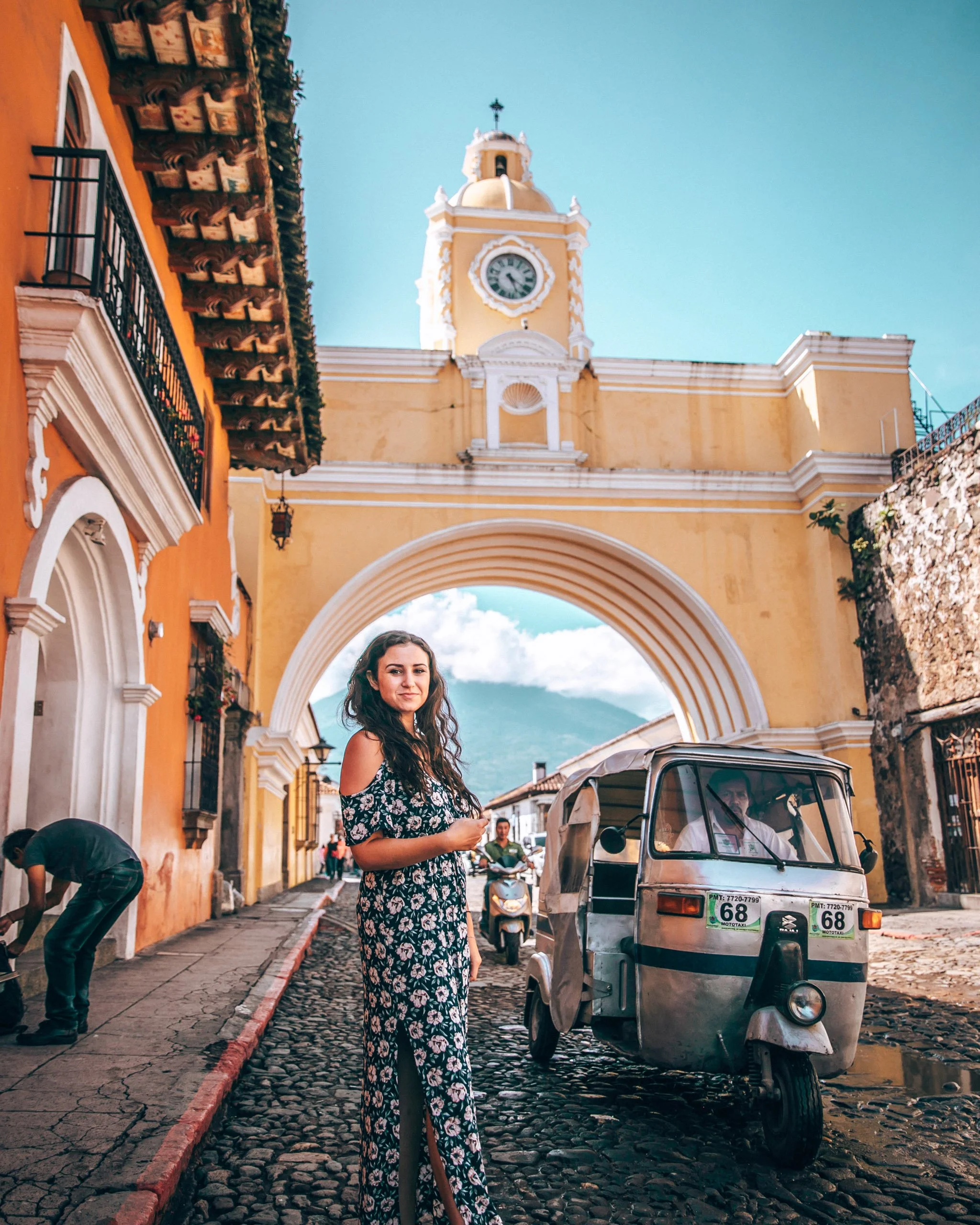 A young woman in a floral dress stands on a cobblestone street in front of a three-wheeled auto rickshaw in a historic area with colorful buildings and a clock tower in the background. The buildings are painted in warm colors with balconies, and the 