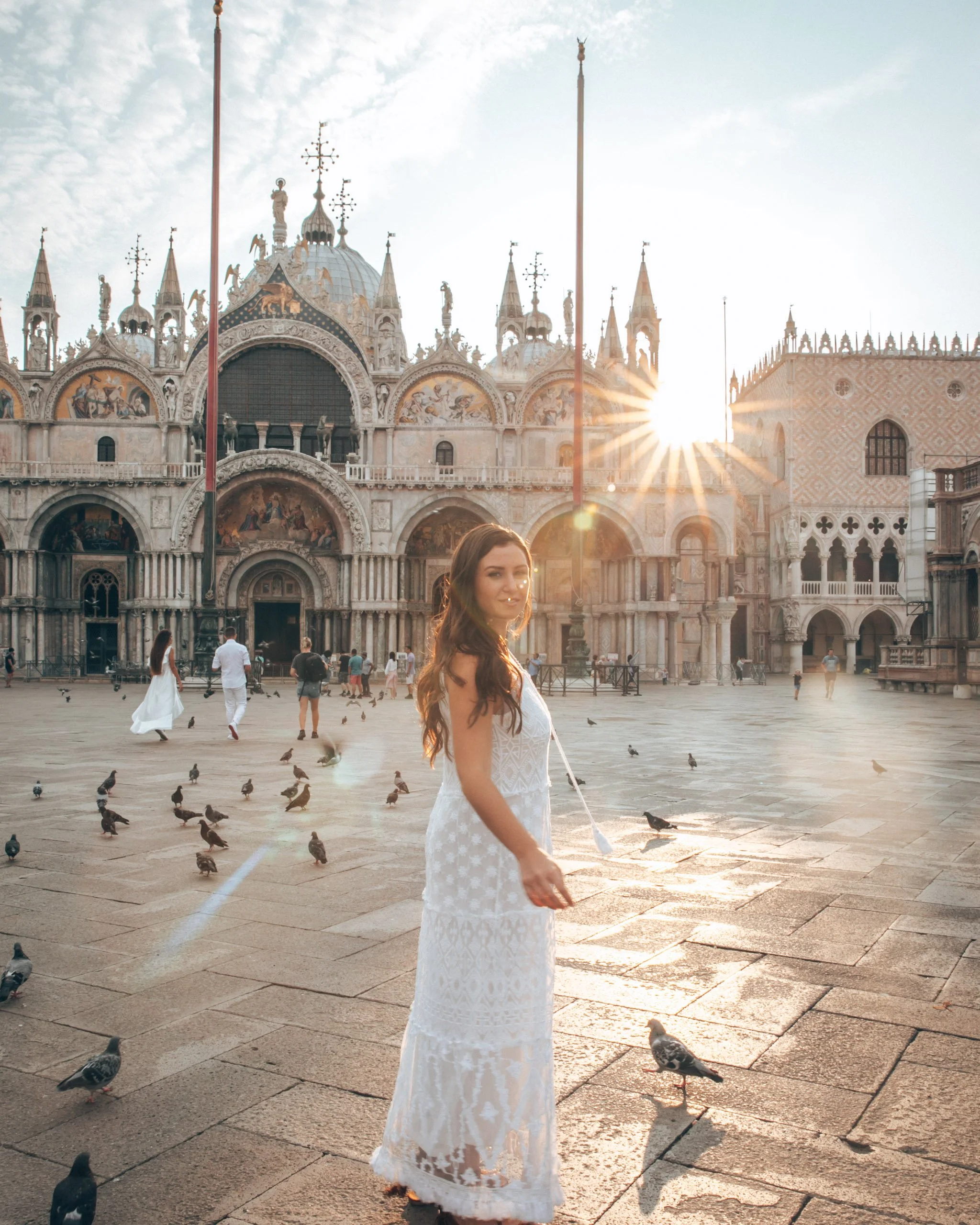 A woman with long brown hair in a white lace dress is standing on a stone square in Venice, Italy, with pigeons around her and the Doge's Palace in the background, illuminated by a setting sun.