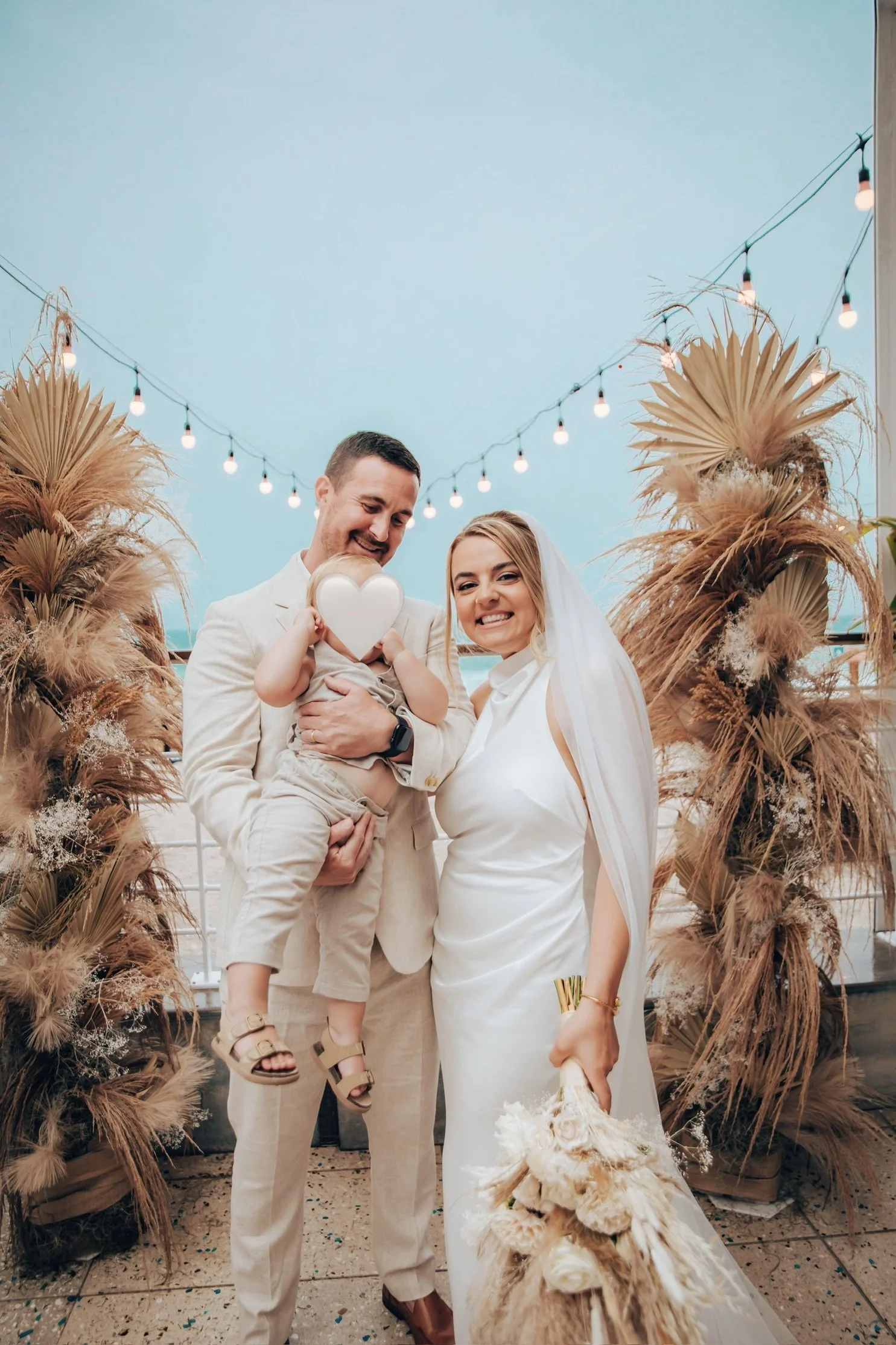 A wedding celebration showing a bride and groom with a young child, standing in front of a decorated backdrop with dried palm leaves and string lights. The bride is holding a bouquet, and the groom is holding the child who is holding a white heart-sh