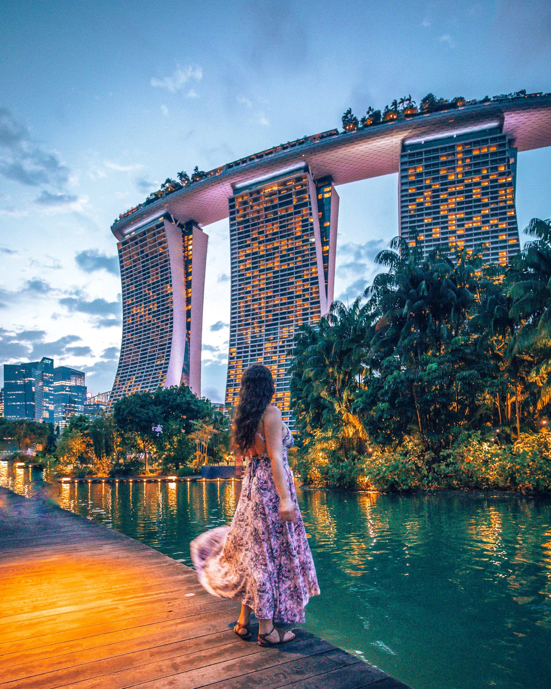A woman with long hair wearing a floral dress and sandals standing on a wooden dock, looking at the water with the Marina Bay Sands hotel in Singapore illuminated behind her during dusk.