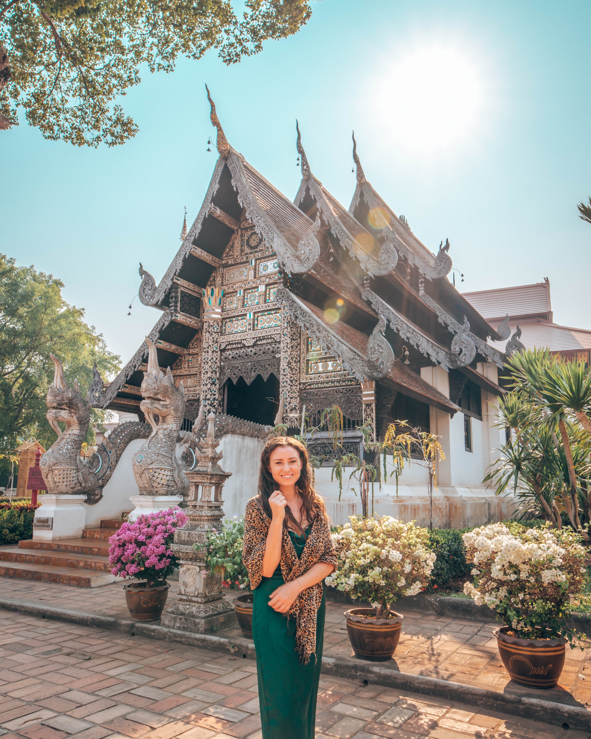 A woman standing outdoors in front of a traditional Thai temple, smiling, with potted flowers and trees around her, during bright daylight.
