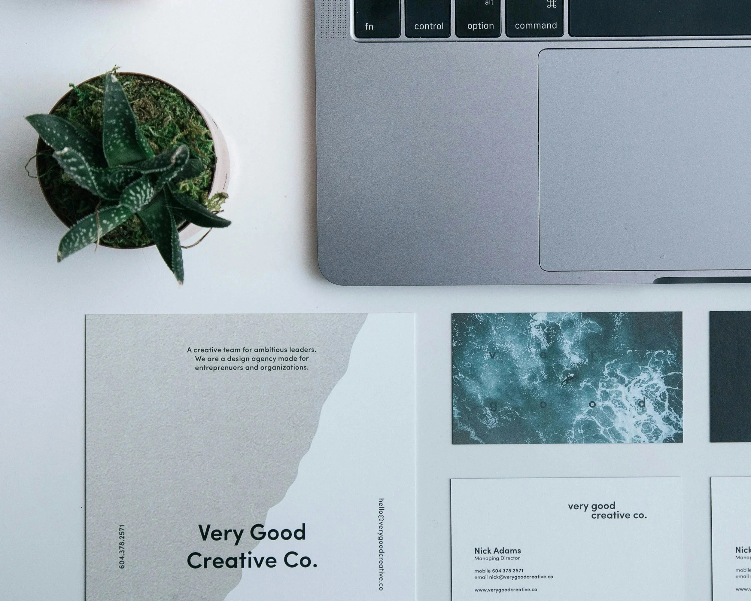 A magazine on a white surface next to a laptop and a potted plant, with business cards or flyers featuring the name "Very Good Creative Co." and contact information.