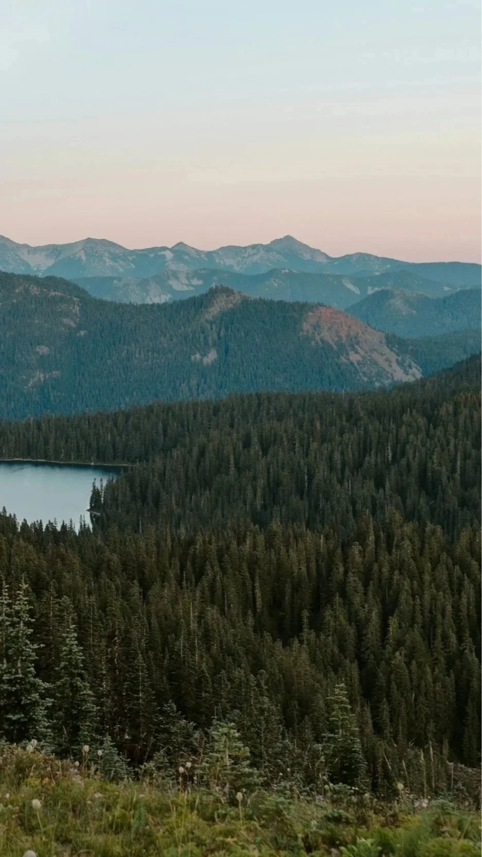 A scenic view of a mountain landscape with a lake surrounded by dense evergreen forests, layered mountains in the background, and a colorful sky at either sunrise or sunset.