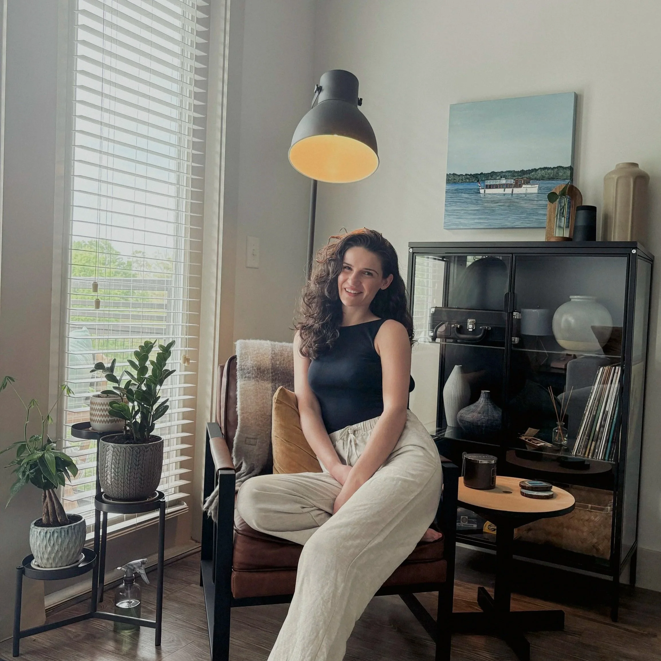 Brooke Eastgate owner of BE Landscape Artist Inc. sitting on a brown armchair in a living room, smiling, with a black sleeveless top and beige pants, surrounded by houseplants and modern decor.