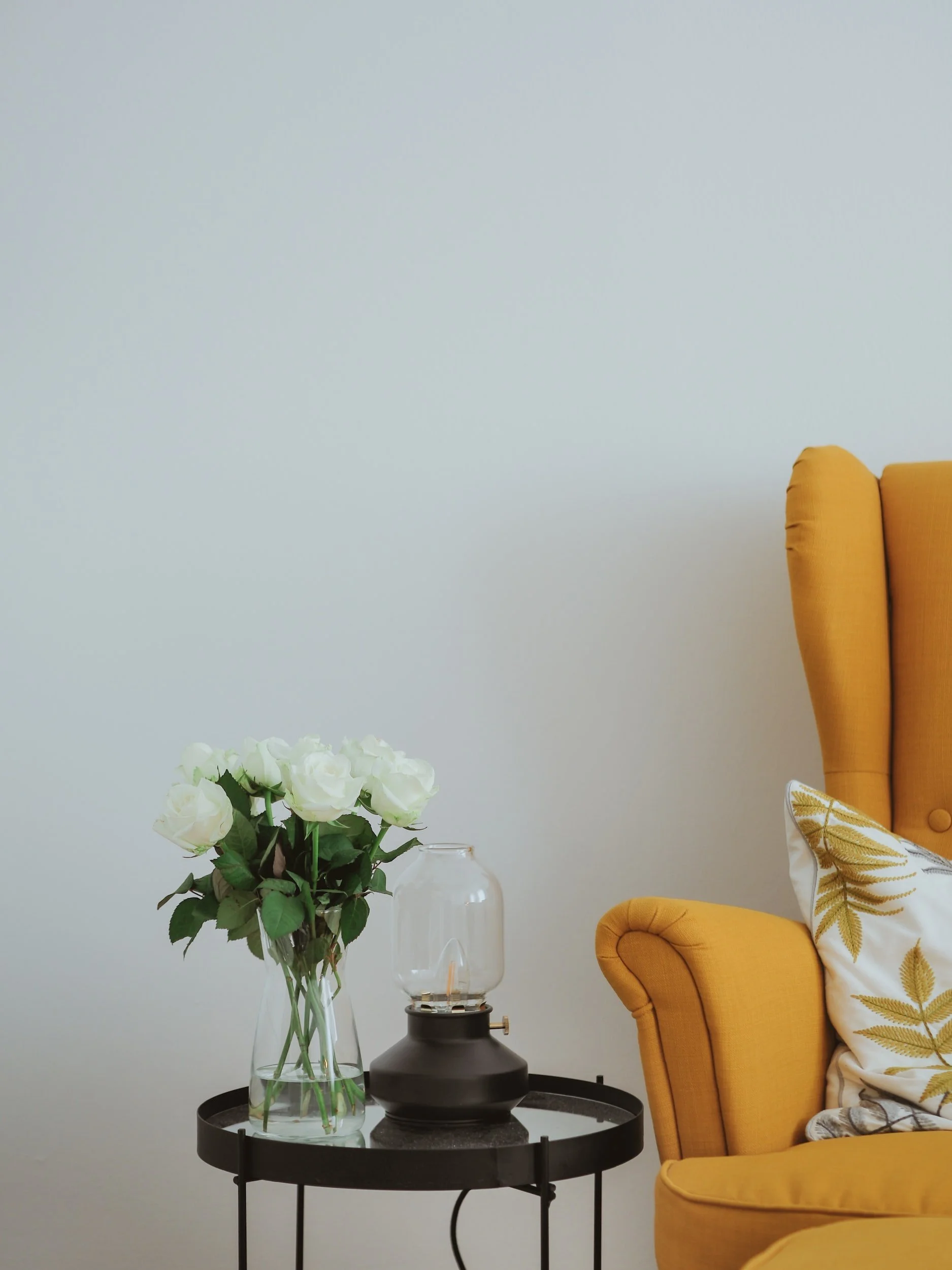 Yellow armchair with leaf-patterned pillow and a small black round side table holding a clear glass vase with white roses and a lamp with a glass cover.