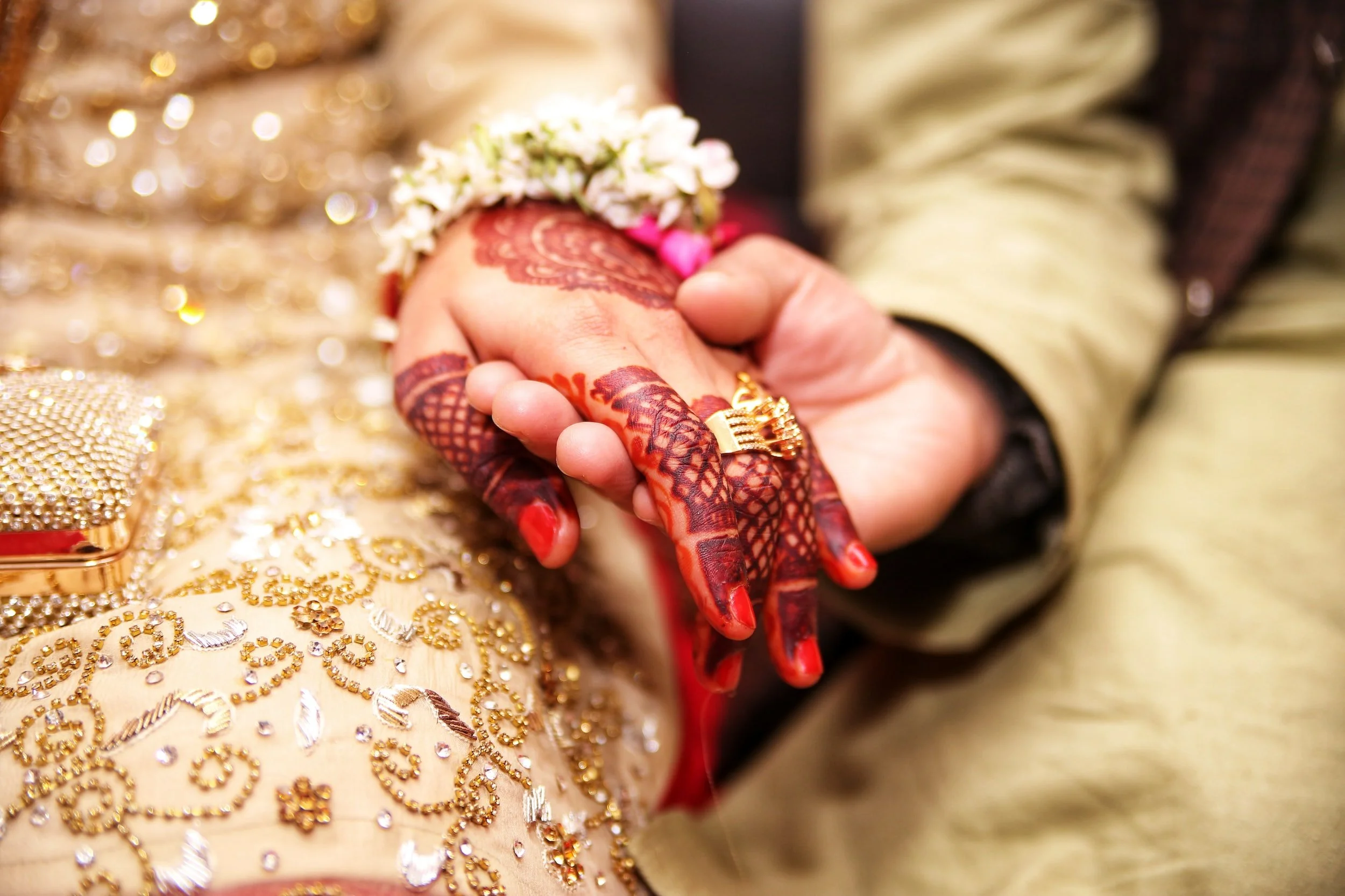 Close-up of a bride and groom holding hands, with the bride's hand adorned with henna designs, rings, and jewelry, including a floral bracelet, and the groom wearing a traditional outfit with gold embroidery.
