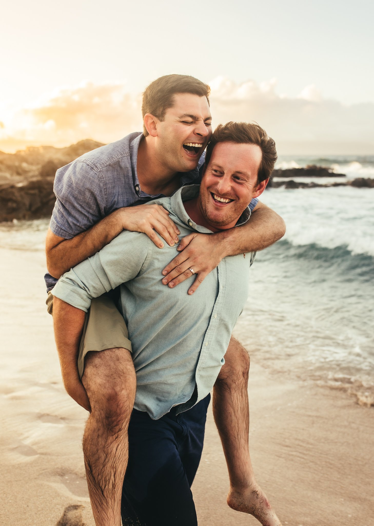 two men laughing as one man is giving the other a piggy back ride on the beach at ironwoods beach in kapalua maui