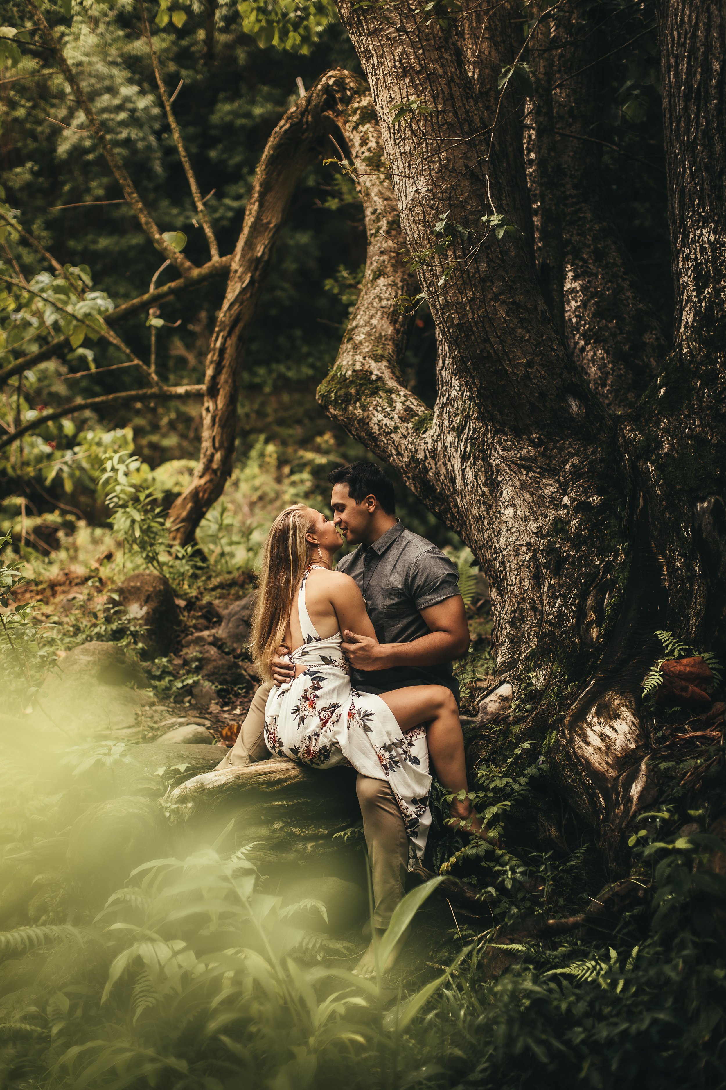 woman and man sitting on log, facing eachother kissing immersed in lush jungle at iao valley in maui hawaii