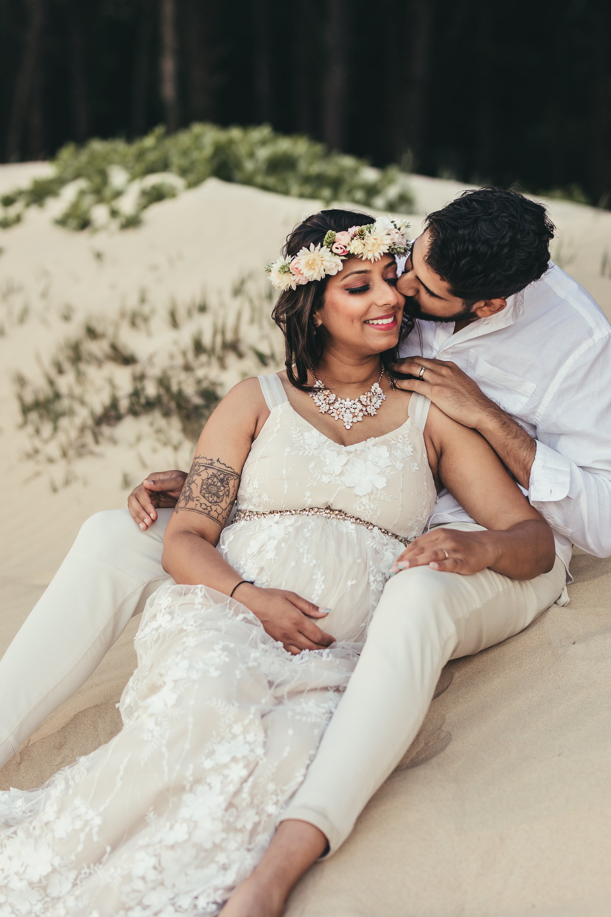 pregnant indian woman wearing flower crown sitting in between man's legs on beach while he kisses her cheek in maui hawaii