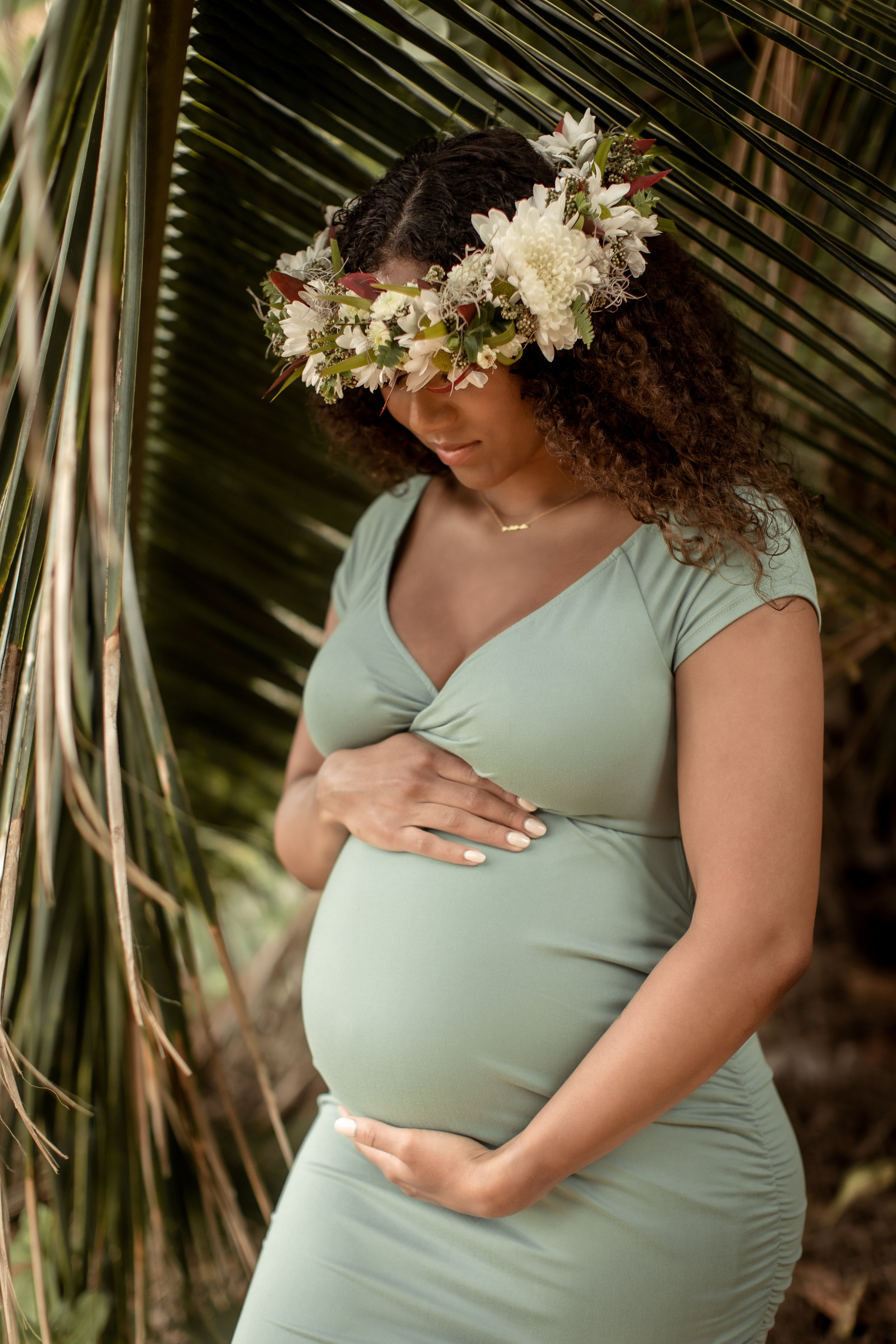 pregnant afican american woman holding tummy wearing flower crown in light green dress surrounded by palm leaves