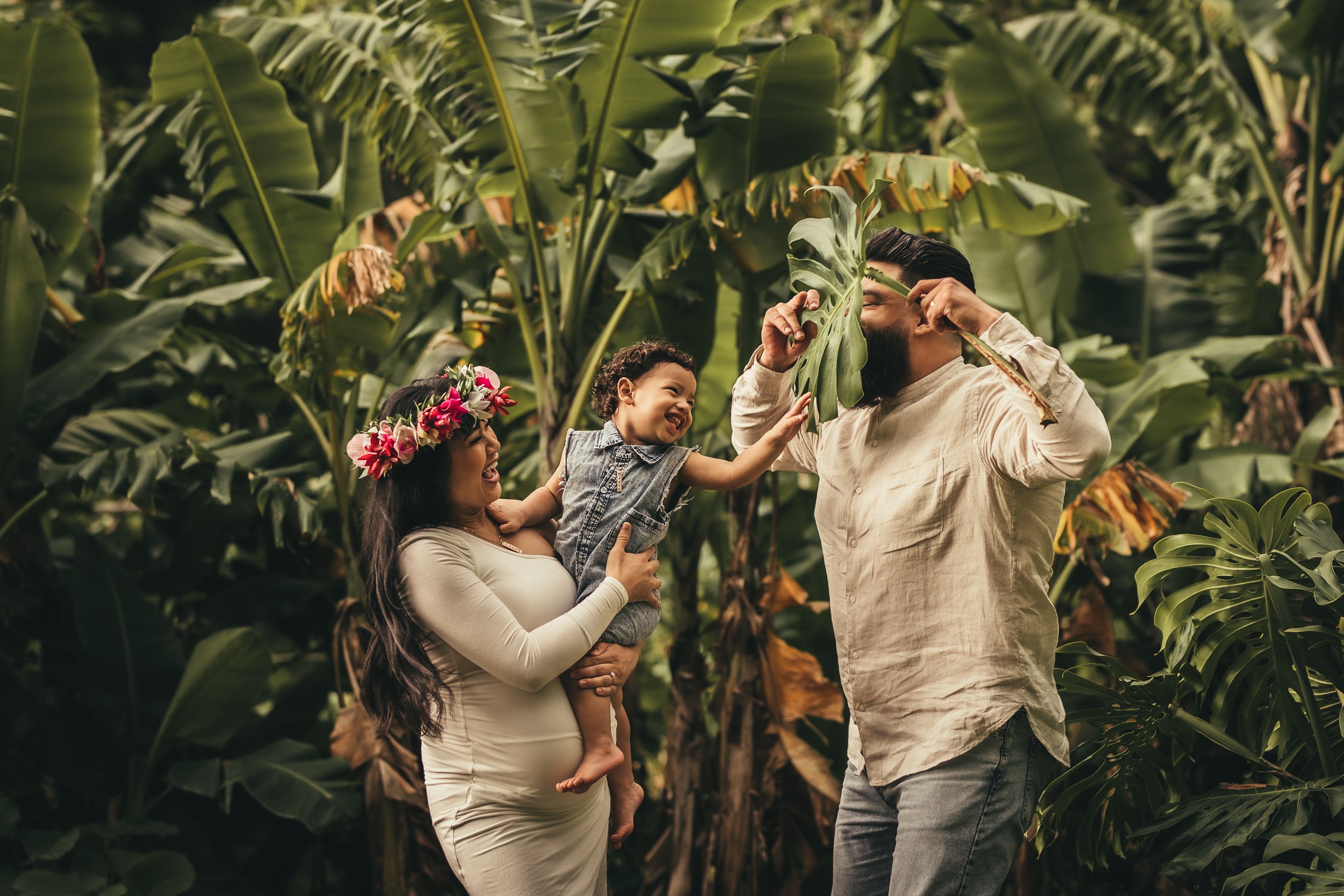 pregnant mother, father and toddler son in mom's arms playing with jungle leaves in iao valley, maui hawaii