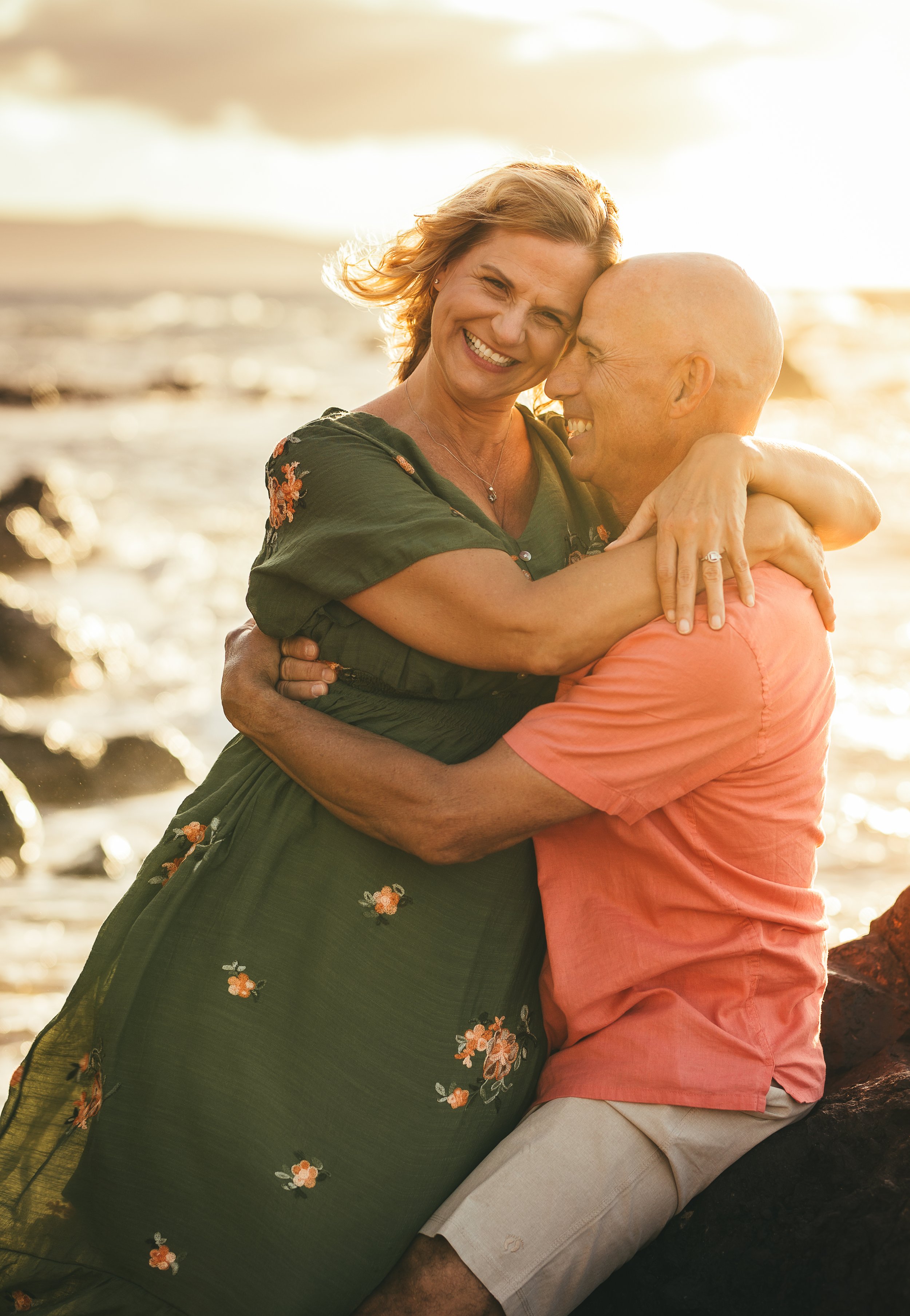 woman embracing man sitting on rock while she looks at camera smiling at sunset on maui hawaii
