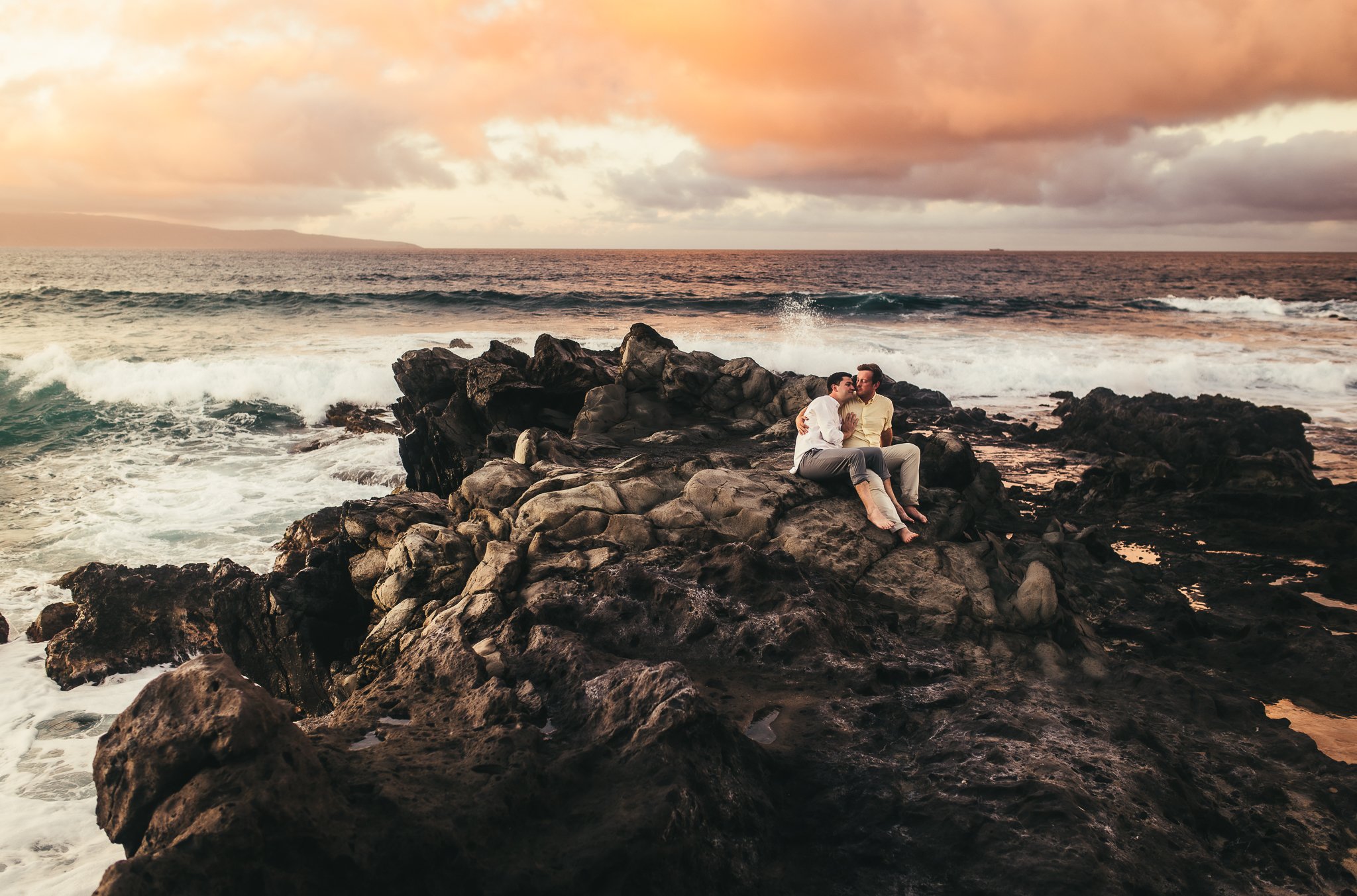 two men embracing eachother on lava rocks while the large ocean crashes behind them at ironwoods beach in kapalua maui