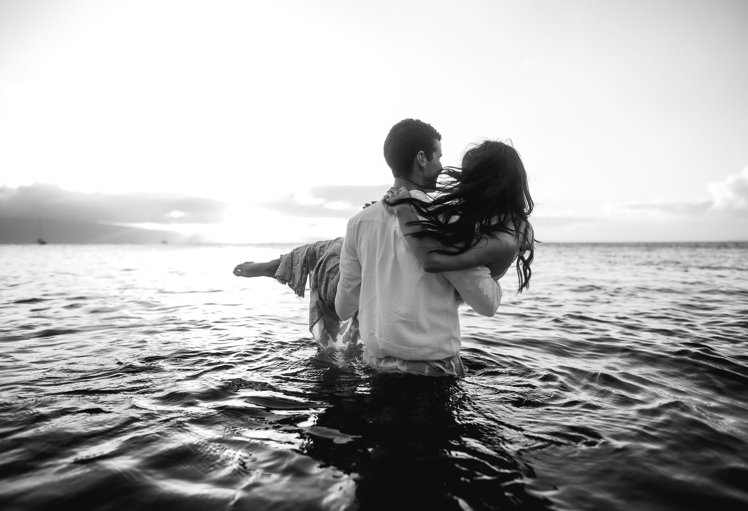 black and white photo of back view of man carrying woman into the ocean with wind blowing her hair