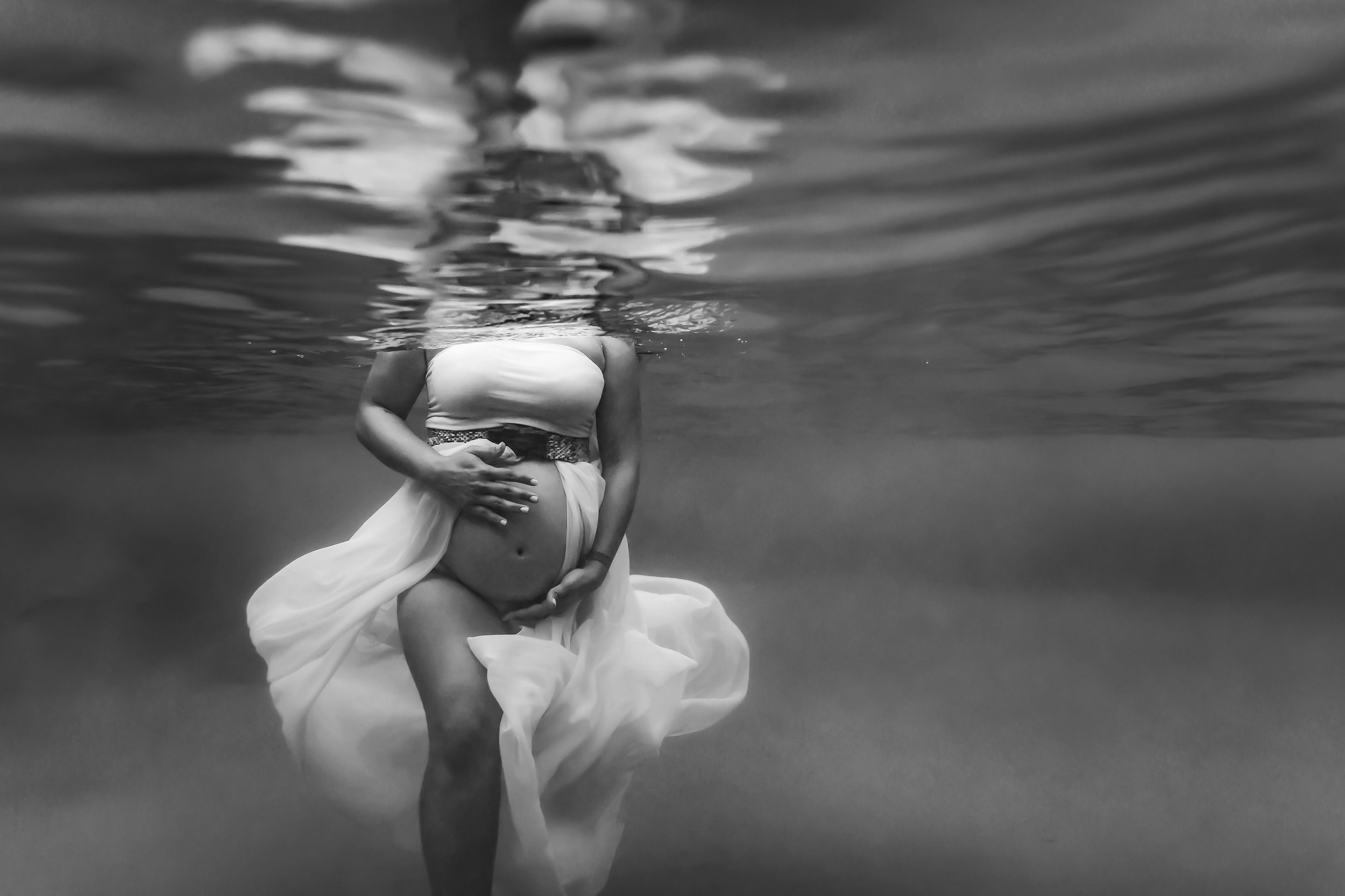 black and white photo of pregnant woman holding tummy underwater