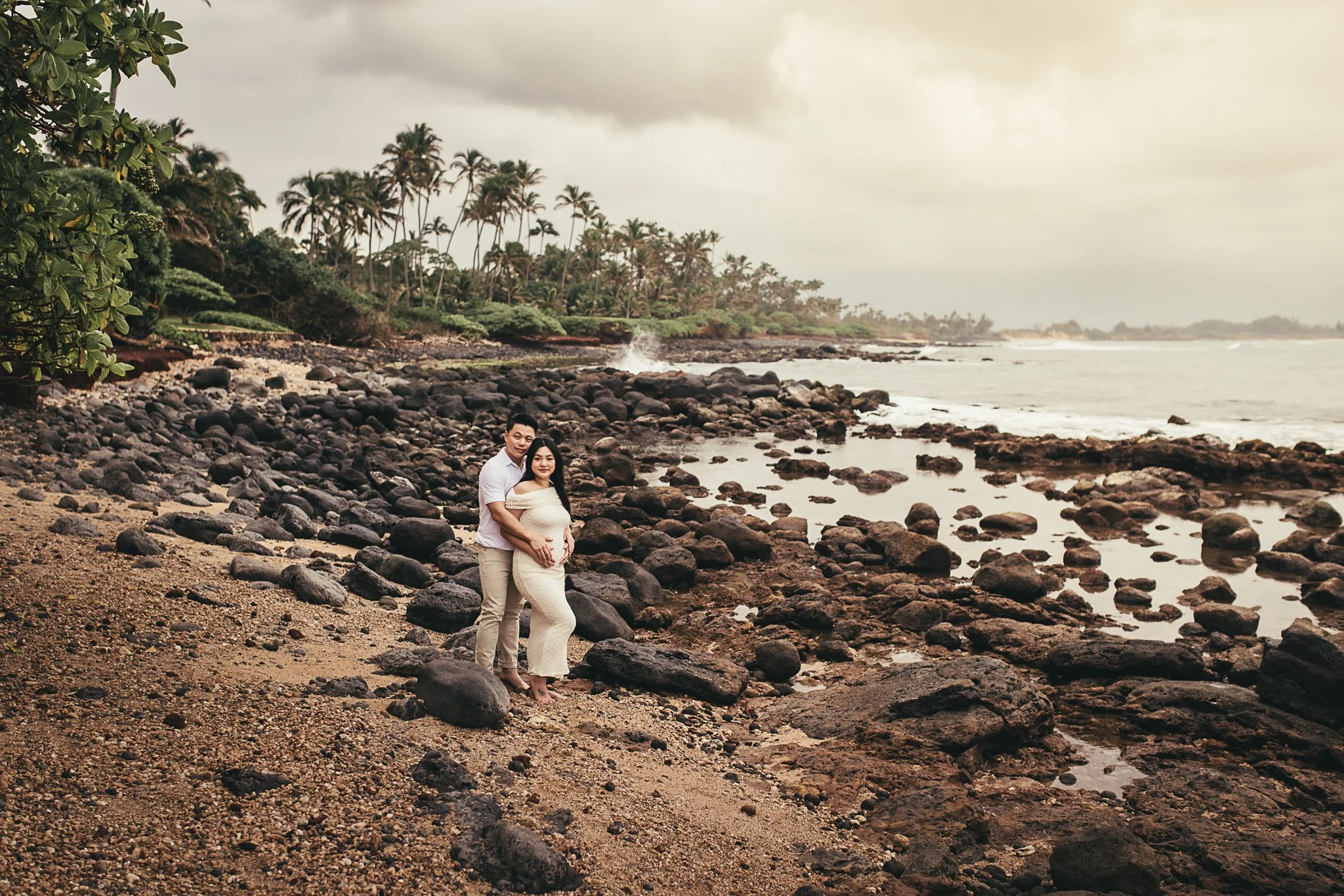 pregnant woman in white dress with man in white shirt embracing each other in lava rock shoreline with palm trees in background on maui at sunset