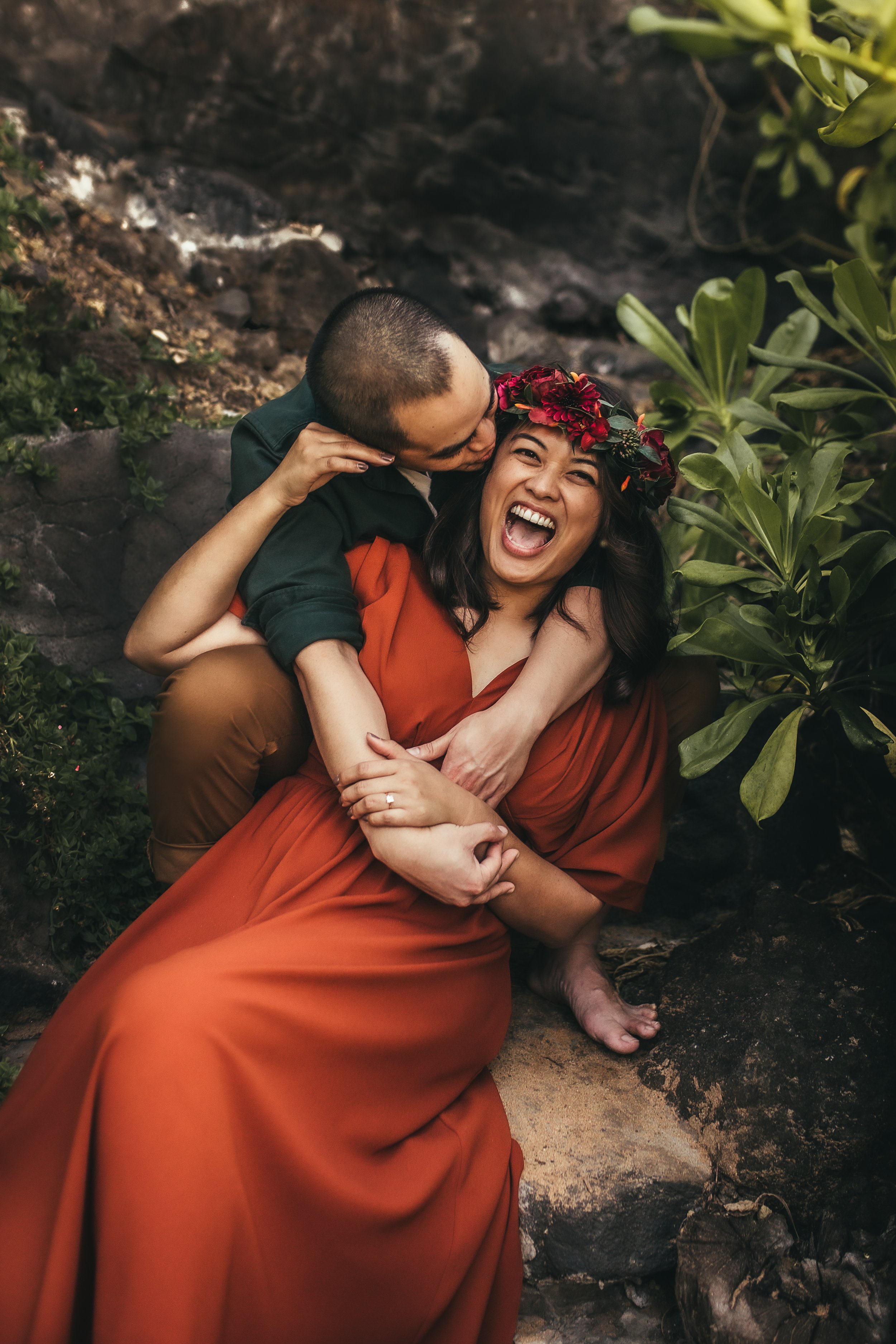 woman in red dress wearing flower crown laughing as she sits in between mans legs and he kisses her cheek in maui hawaii