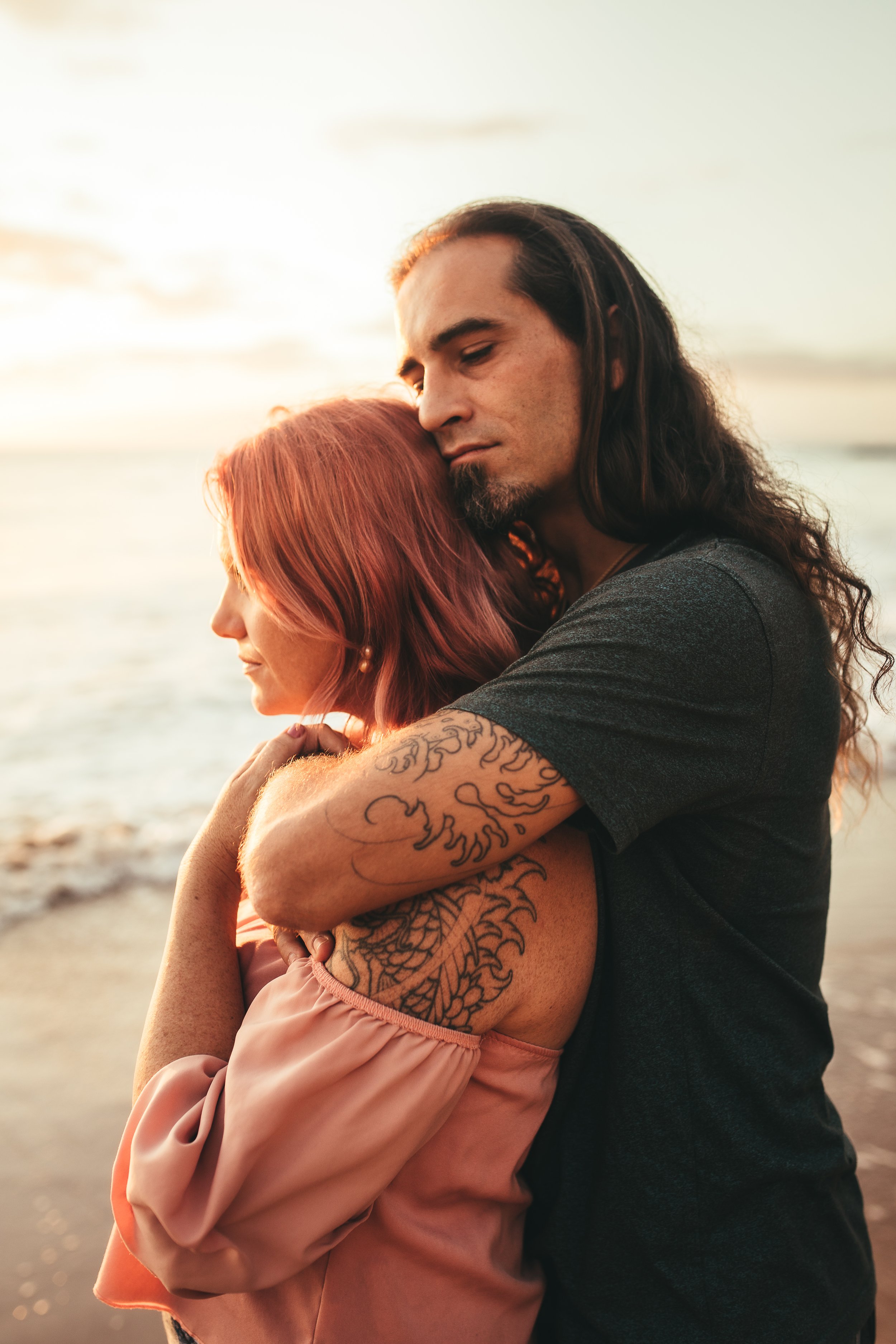 man embracing woman from behind with his eyes closed while she gazes into ocean