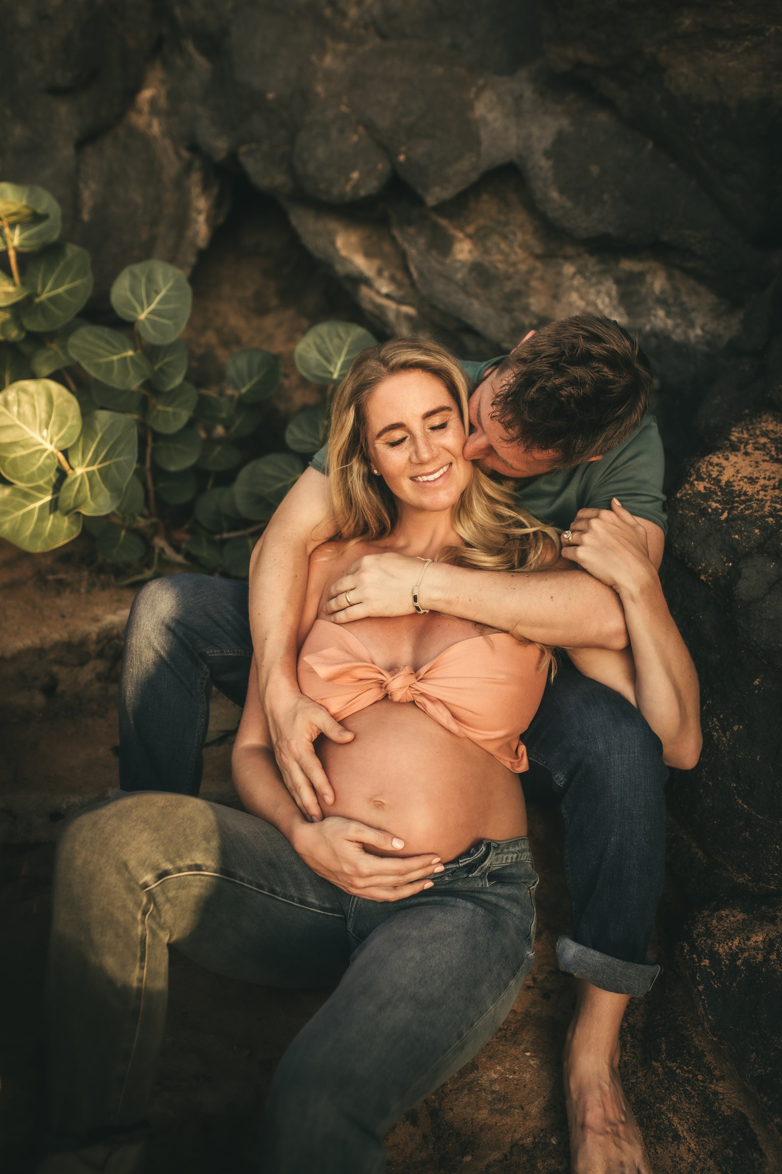 pregnant woman sitting in between mans legs on steps while he kisses her cheek in maui hawaii