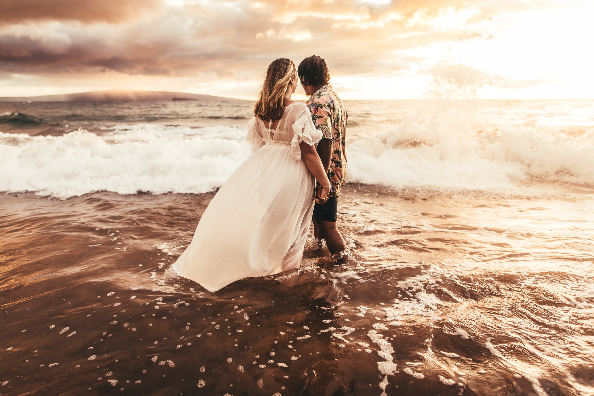 man and woman holding hands while standing in ocean admiring sunset and island of kahoolawe in maui hawaii