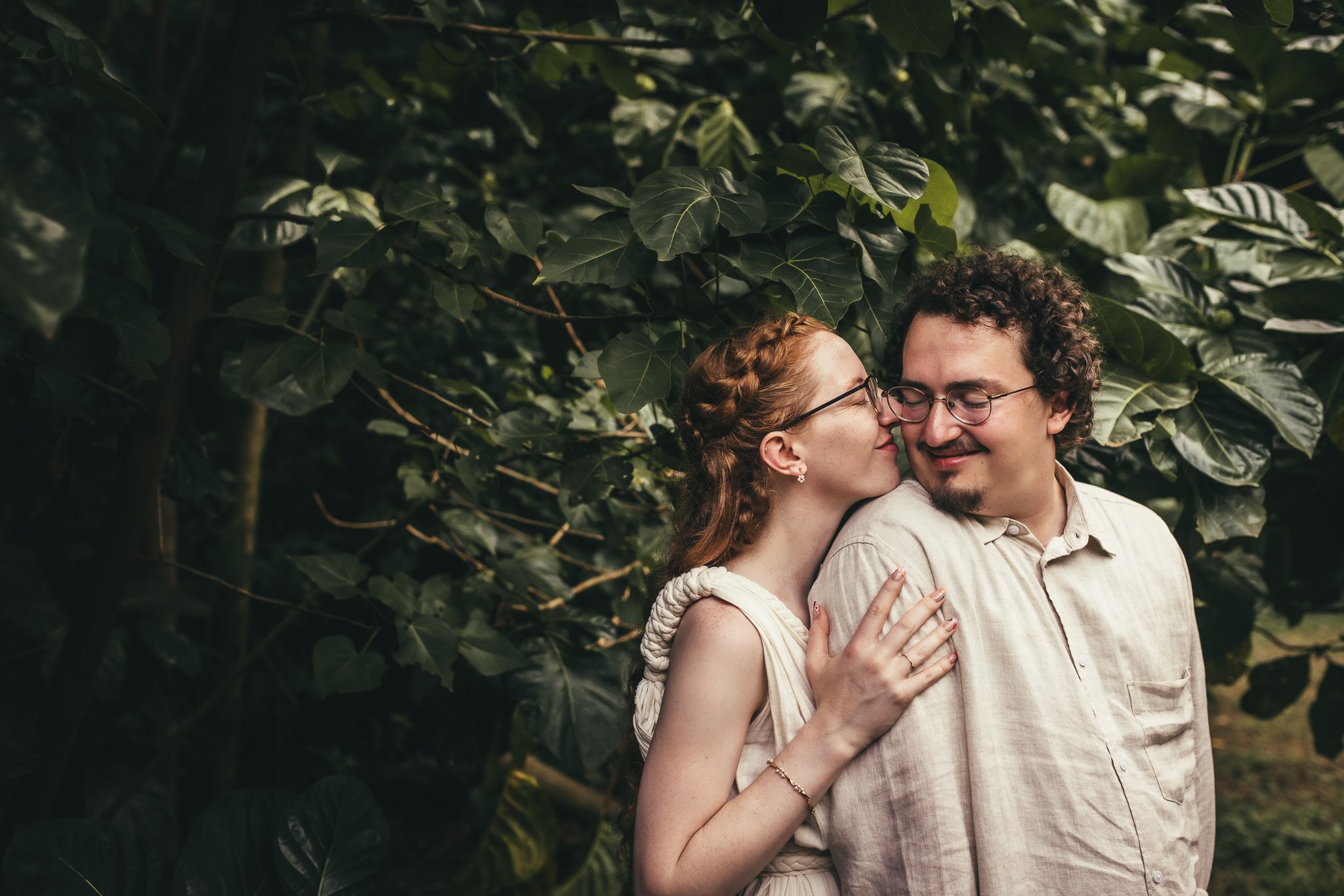 woman standing behind man, embracing him and touching her nose to his ear as he looks over his shoulder in lush jungle in maui hawaii