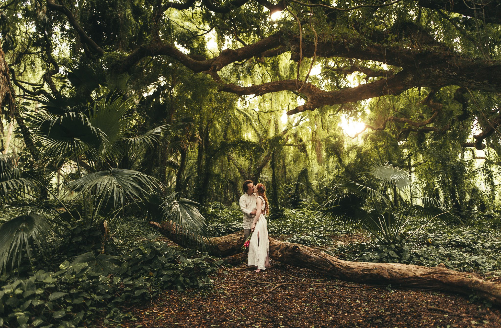 man and woman leaning on large log while gazing into eachother's eyes in lush green jungle at honolua bay in maui hawaii