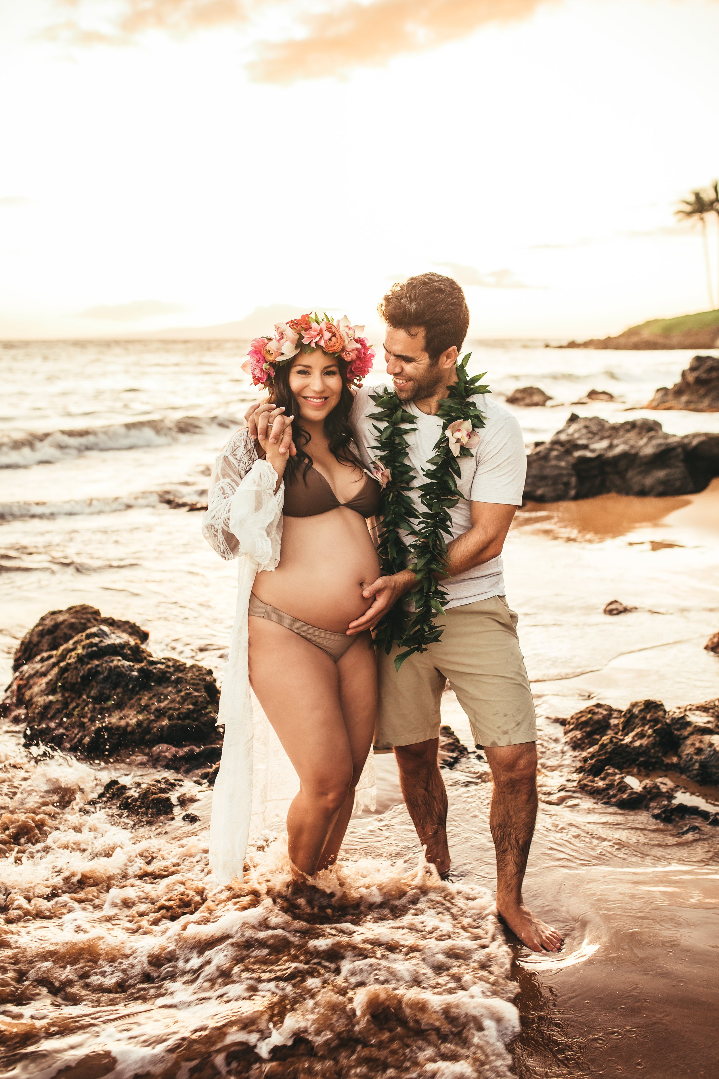 pregnant woman in bikini with white coverup smiling with flower crown and man with hand on her tummy admiring her at sunset on maui hawaii