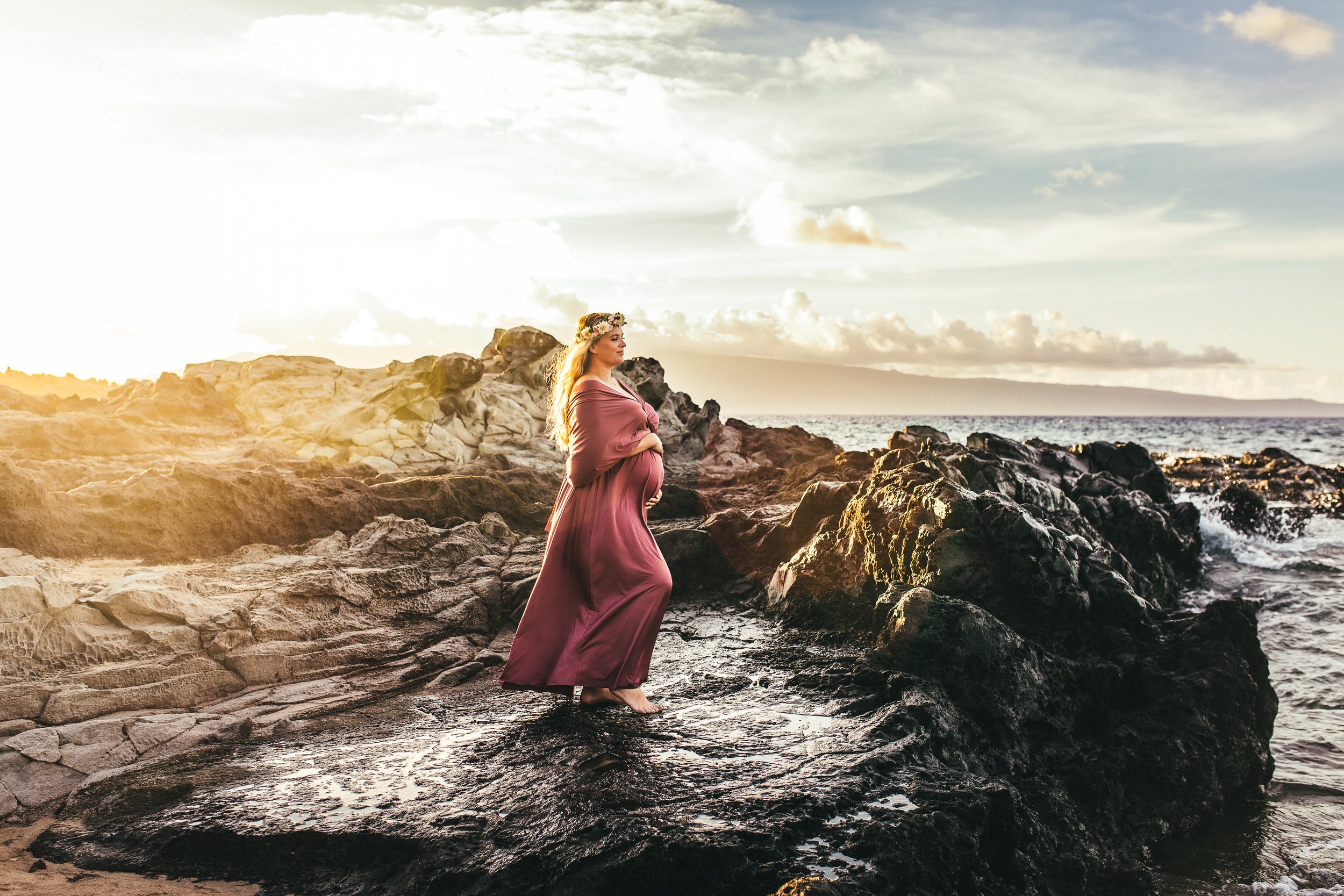 pregnant woman in flowy mauve dress standing cliffside on lava rocks at ironwoods beach in maui hawaii at sunset