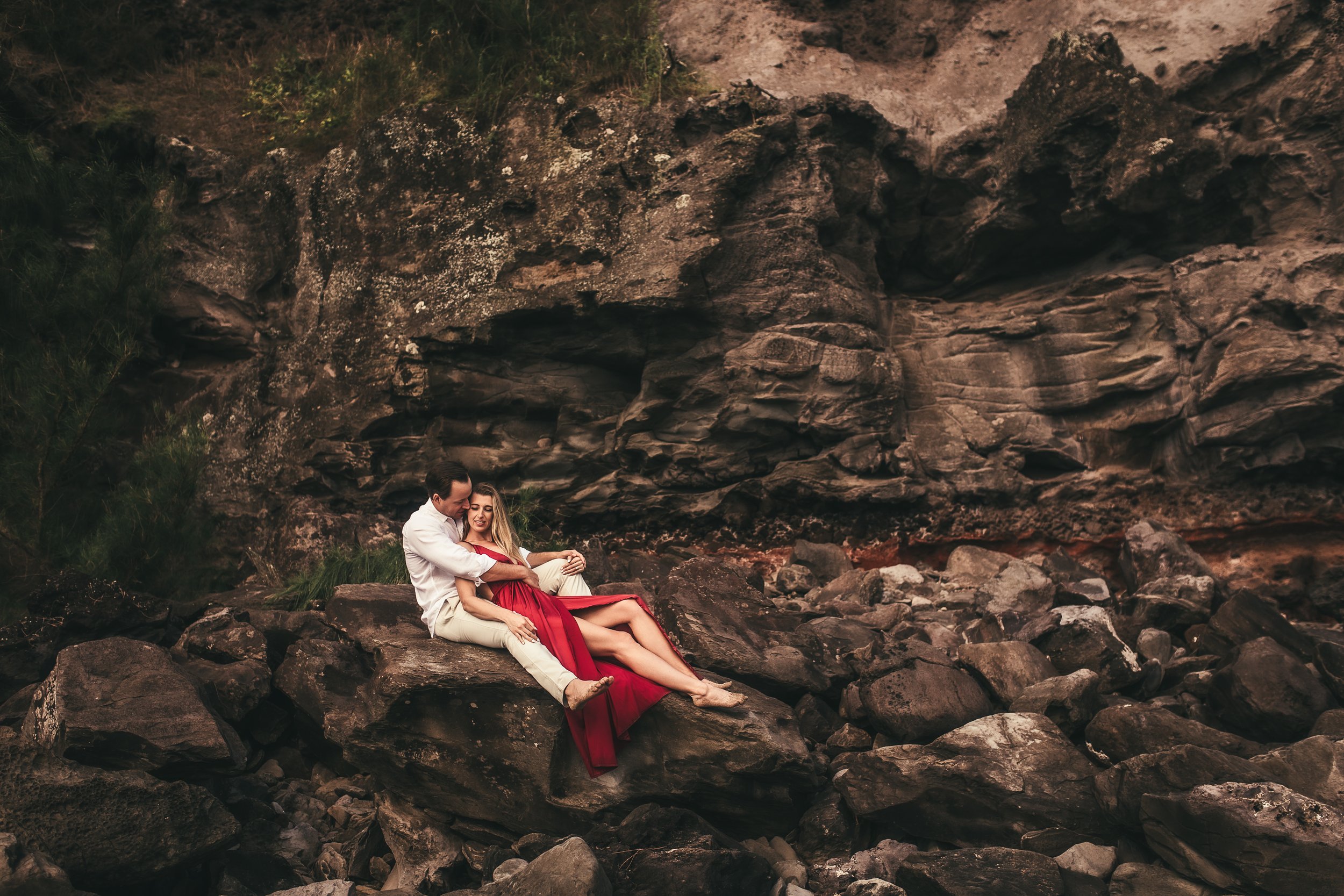 woman sitting in between mans legs as they sit on large lava rock, surrounded by large lava rocks in maui hawaii