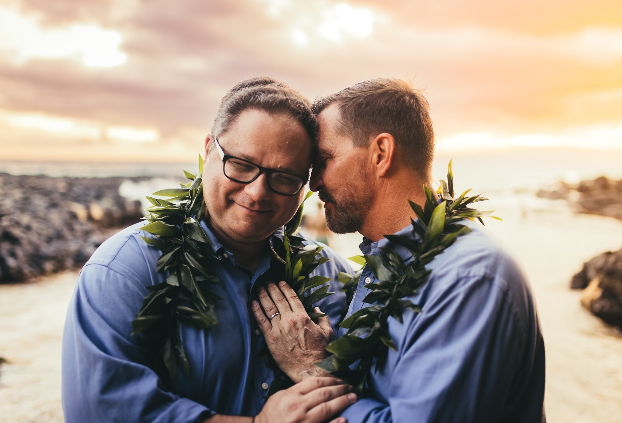 two older men cuddling close together with ti leaf lei around their necks at sunset at secret beach in maui hawaii