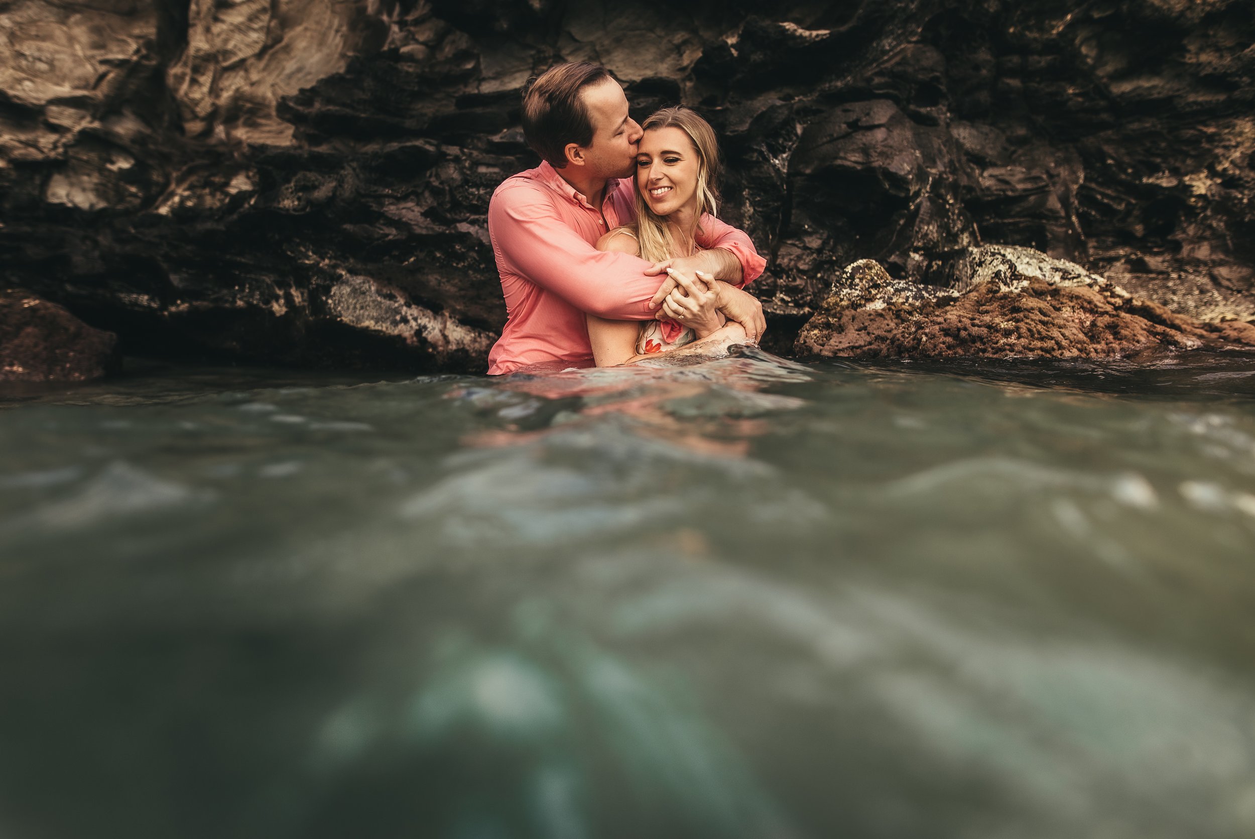 man standing behind woman, embracing her and kissing her head while they are chest deep in the ocean surrounded by lava rocks