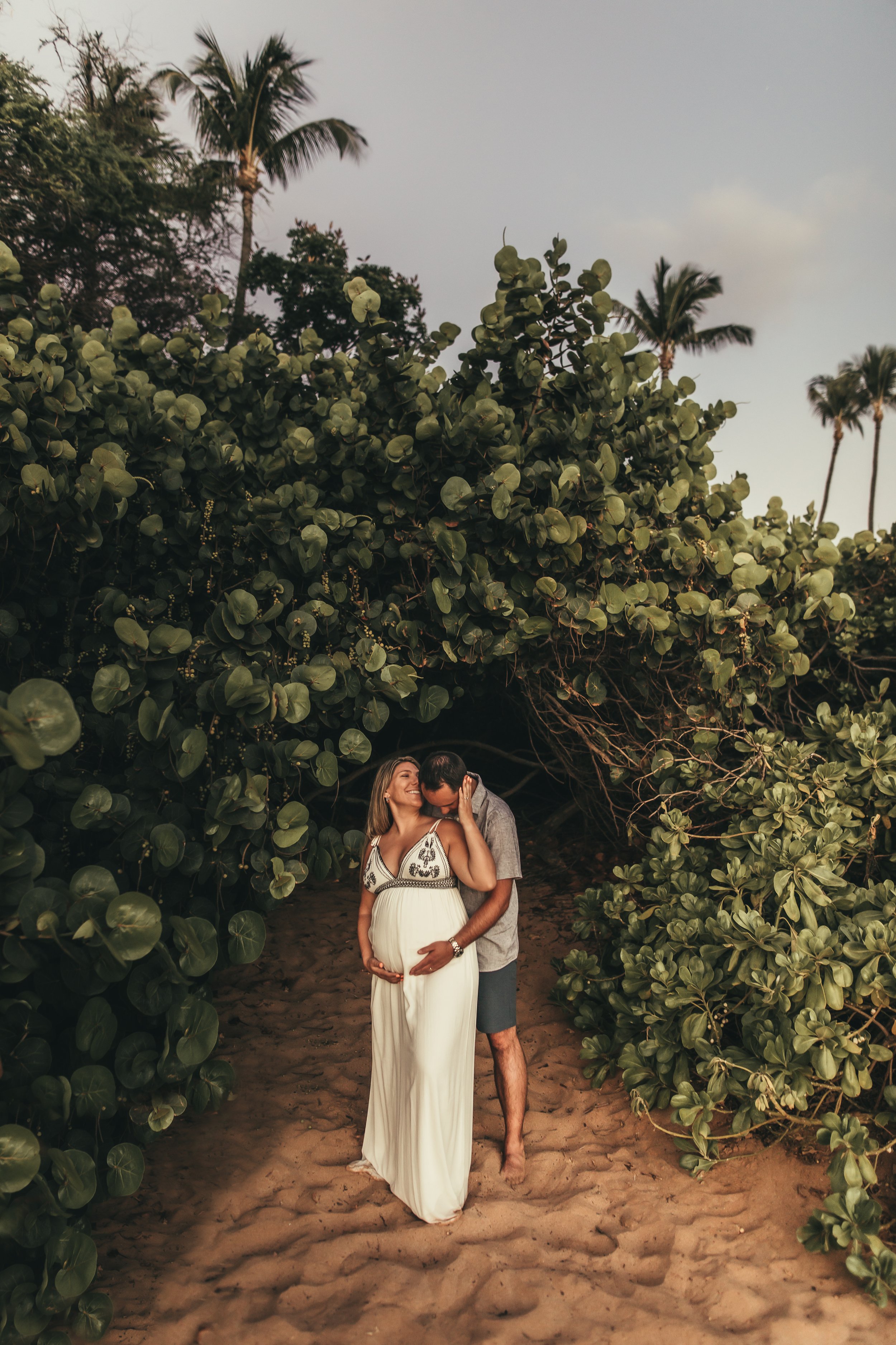 man standing behind pregnant woman with hands on her tummy on a beach in maui hawaii